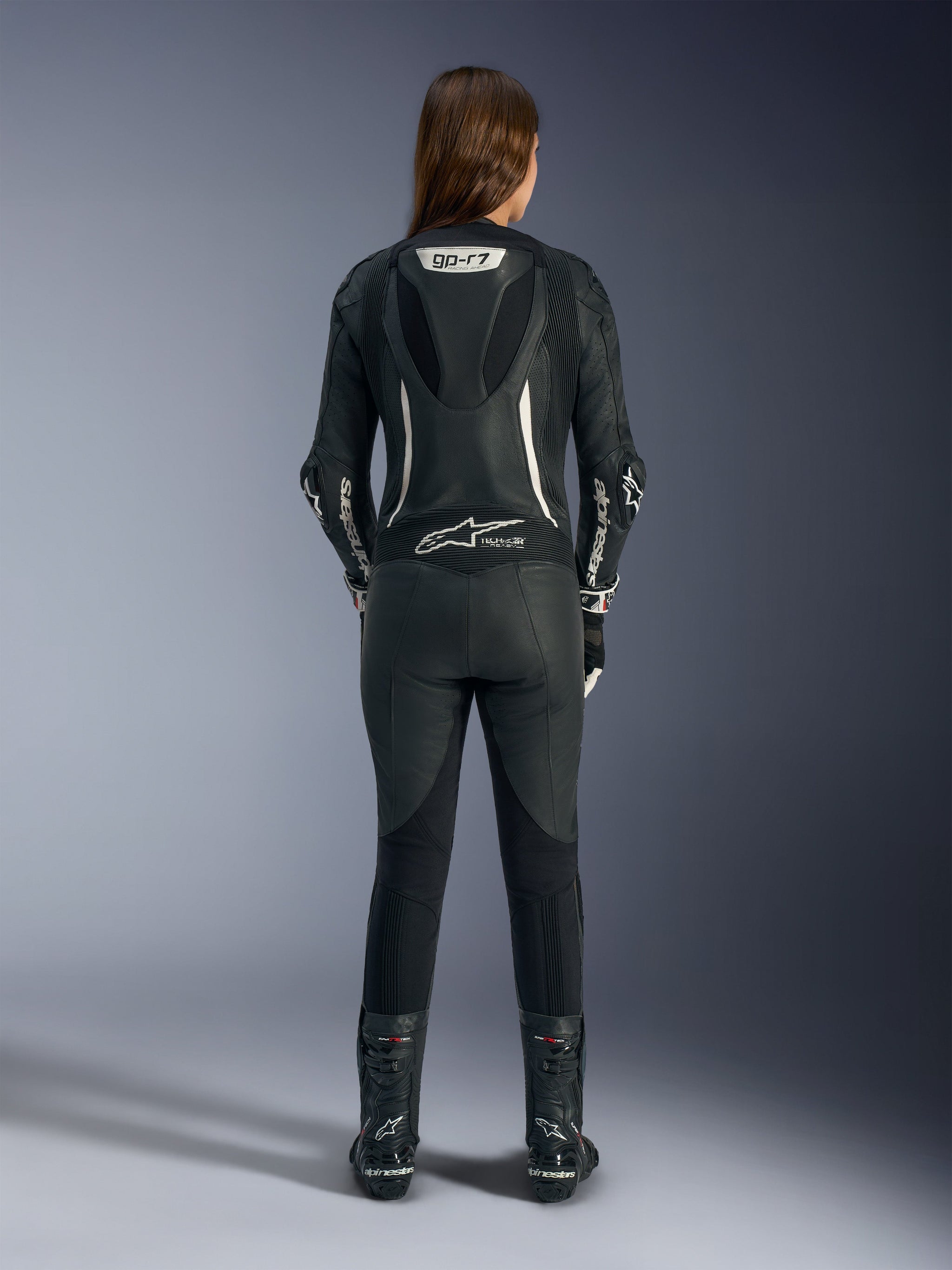 Female rider shown from the back wearing the Alpinestars Stella GP-R7 1-Piece Leather Suit, black leather with white branding, aerodynamic speed hump with GP-R7 logo, Tech-Air Ready designation, paired with black racing boots, professional motorcycle racing gear optimized for women