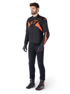Alpinestars T-Jaws V4 vandtæt jakke, sort og rød fluo, båret af en mandlig model med sorte bukser, handsker og støvler, med klare orange detaljer og Alpinestars-logoet, sporty design til motorcykelkørsel