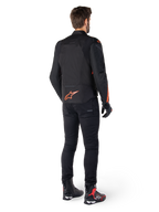 Alpinestars T-Jaws V4 vandtæt jakke, sort og rød fluo, set bagfra på en mandlig kører, med et markant rød fluo logo på ryggen, stylet med sorte kørebukser og tekniske støvler, sportsligt motorcykeludstyr