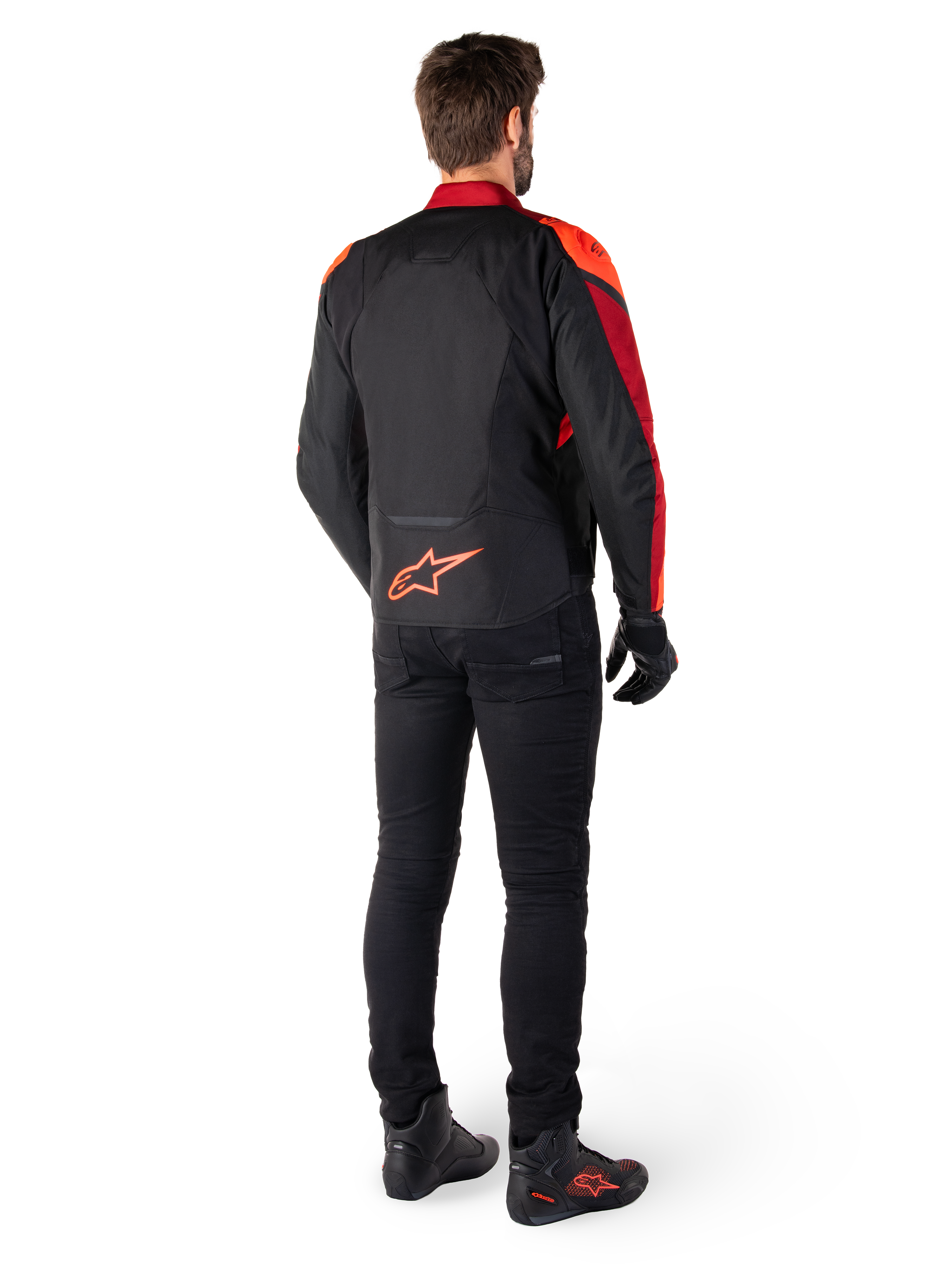 Alpinestars T-Jaws V4 vandtæt jakke i sort, rød fluo og mørkerød, set bagfra på en kører iført tekstiljakken med sorte bukser og kørselstøvler, med et markant rødt Alpinestars-logo nederst på ryggen.