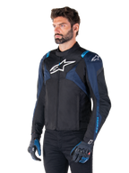Alpinestars T-Jaws V4 Waterproof jakke, motorcykeljakke, sort, mørkeblå og Blithe Blue, båret af en model med matchende handsker og sorte bukser, med hvide Alpinestars-logoer på brystet og skuldrene og blå detaljer på ærmerne, sporty tekstil-køreudstyr