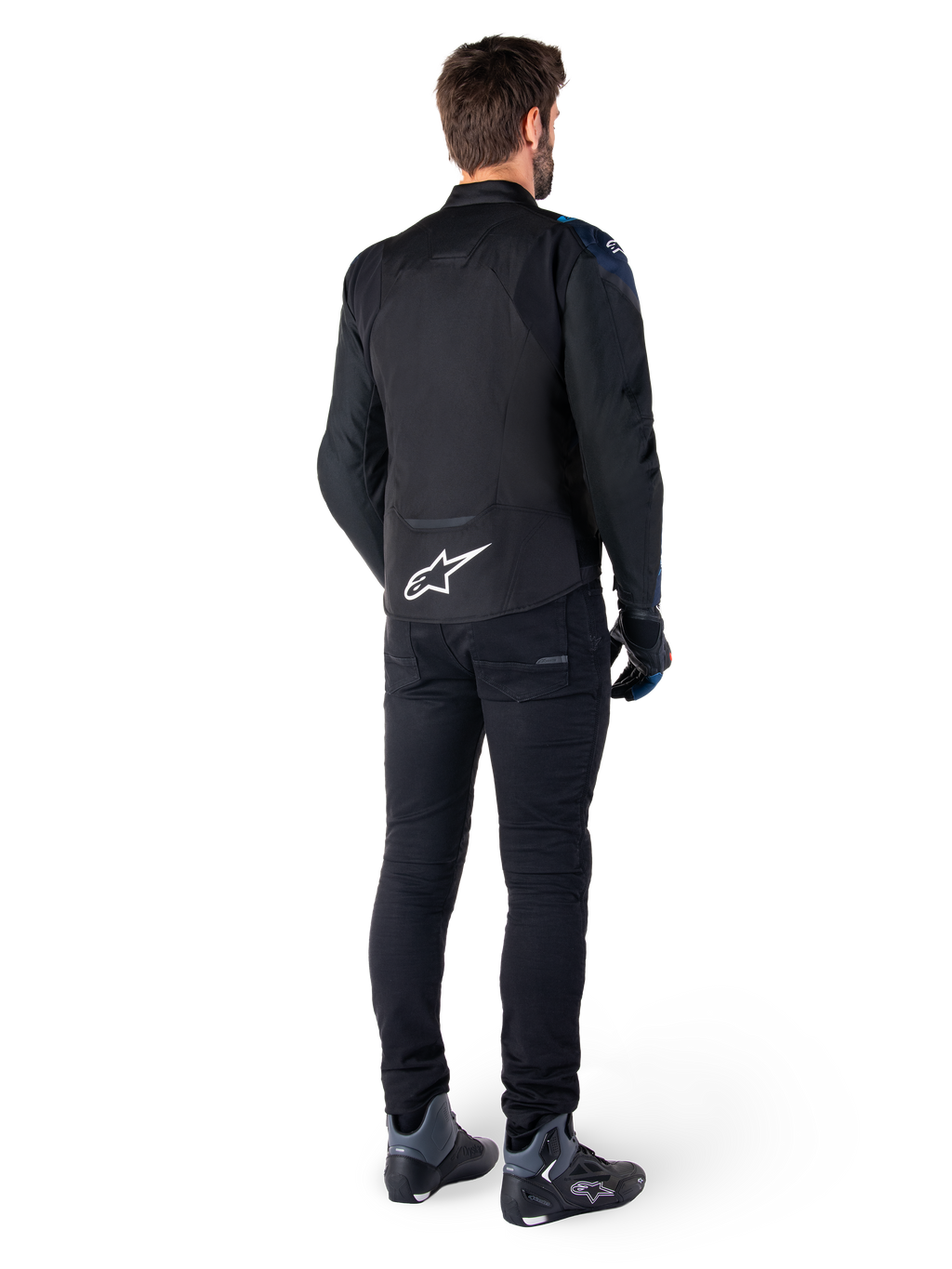 Set bagfra af en mand iført Alpinestars T-Jaws V4 Waterproof Jakke i sort, mørkeblå og Blithe Blue, med et fremtrædende hvidt Astar-logo på lænden, kombineret med sorte bukser og motorcykelsko.