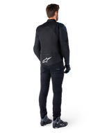 Set bagfra af en mand iført Alpinestars T-Jaws V4 Waterproof Jakke i sort, mørkeblå og Blithe Blue, med et fremtrædende hvidt Astar-logo på lænden, kombineret med sorte bukser og motorcykelsko.