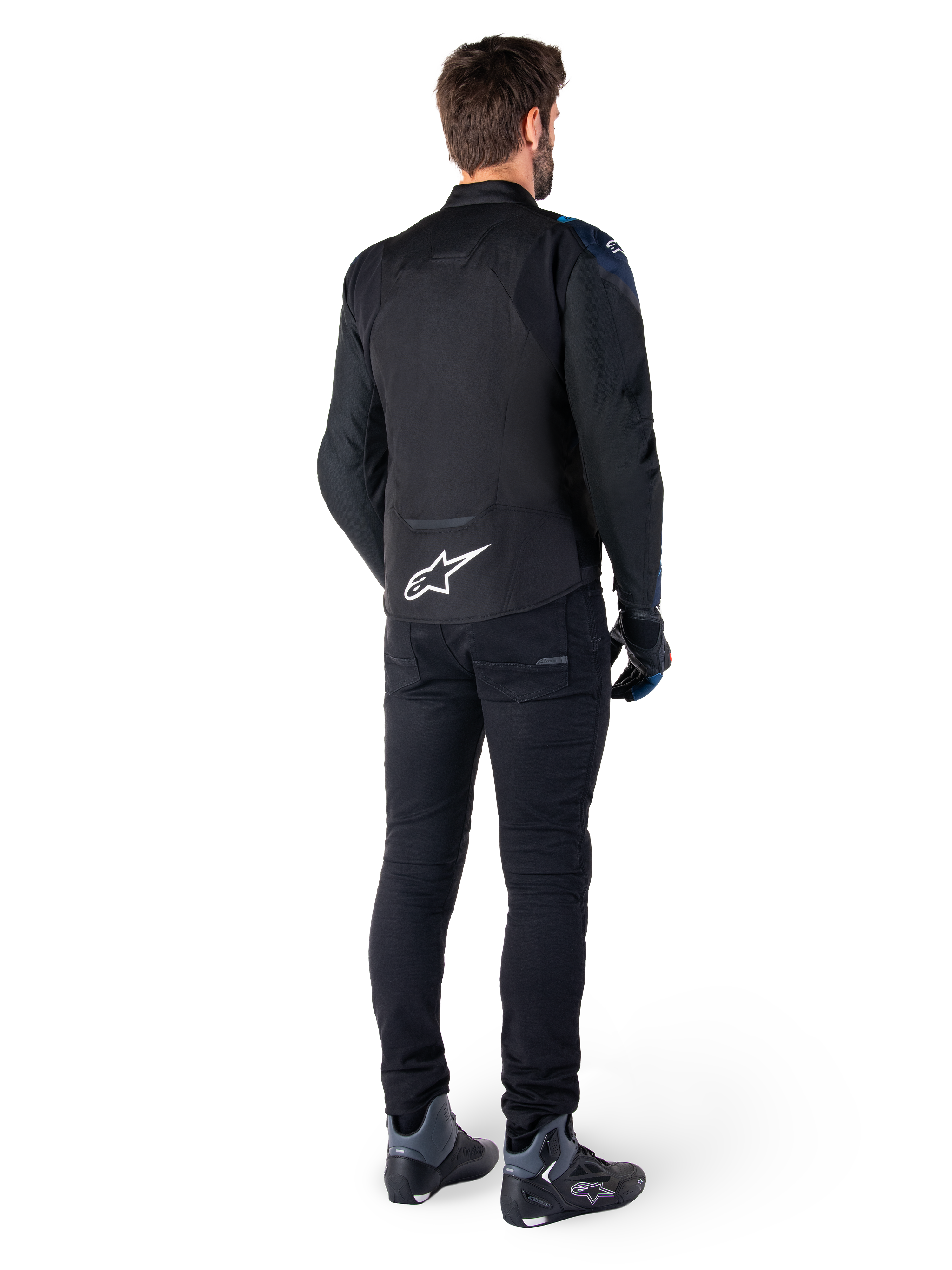 Set bagfra af en mand iført Alpinestars T-Jaws V4 Waterproof Jakke i sort, mørkeblå og Blithe Blue, med et fremtrædende hvidt Astar-logo på lænden, kombineret med sorte bukser og motorcykelsko.