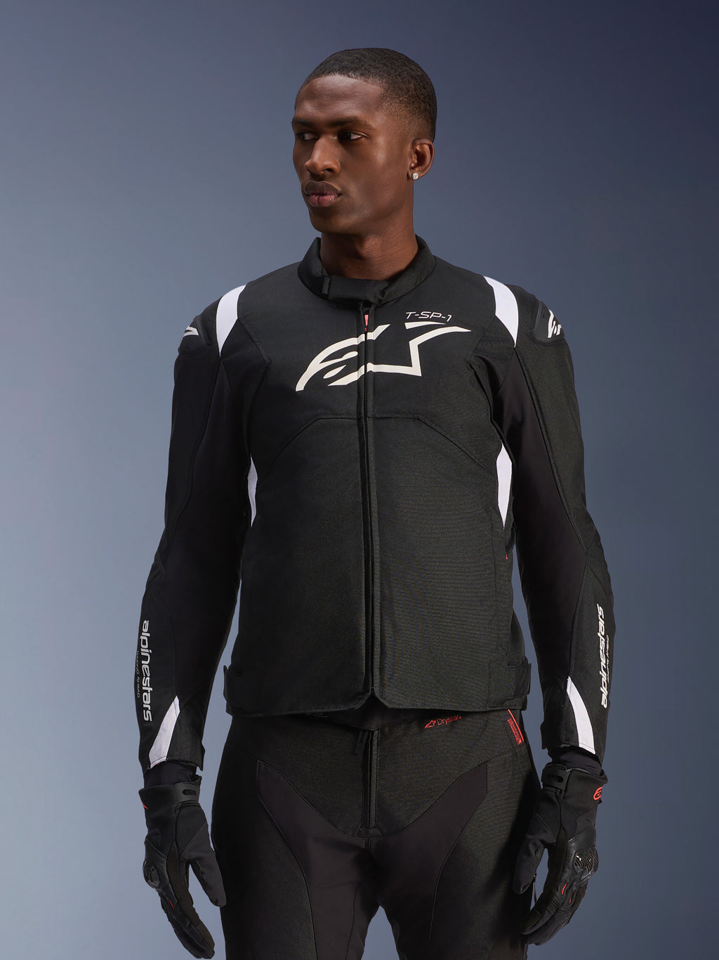 Alpinestars T-SP 1 V2 vandtæt jakke, motorcykeljakke i sportstekstil, sort og hvid med diskrete røde detaljer, båret af en mandlig model med matchende handsker og bukser, med hvidt Alpinestars-logo på brystet, T-SP-1-branding og beskyttende silhuet til landevejskørsel