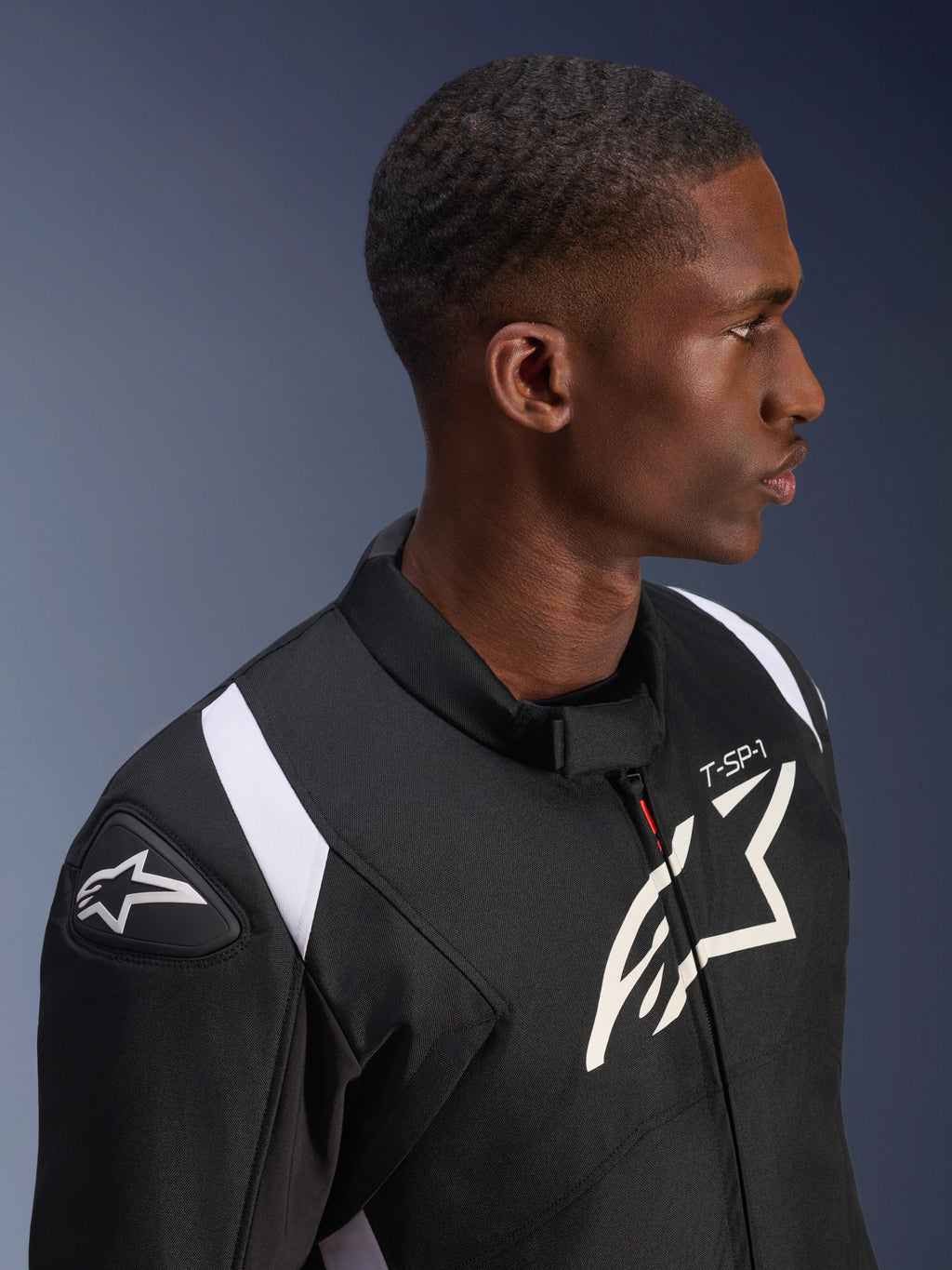 Alpinestars T-SP 1 V2 vandtæt jakke, sort og hvid, profilbillede af en mandlig model iført jakken, Alpinestars-logo på skulderskyder og bryst, 'T-SP-1'-branding, hvide detaljer på skuldrene.