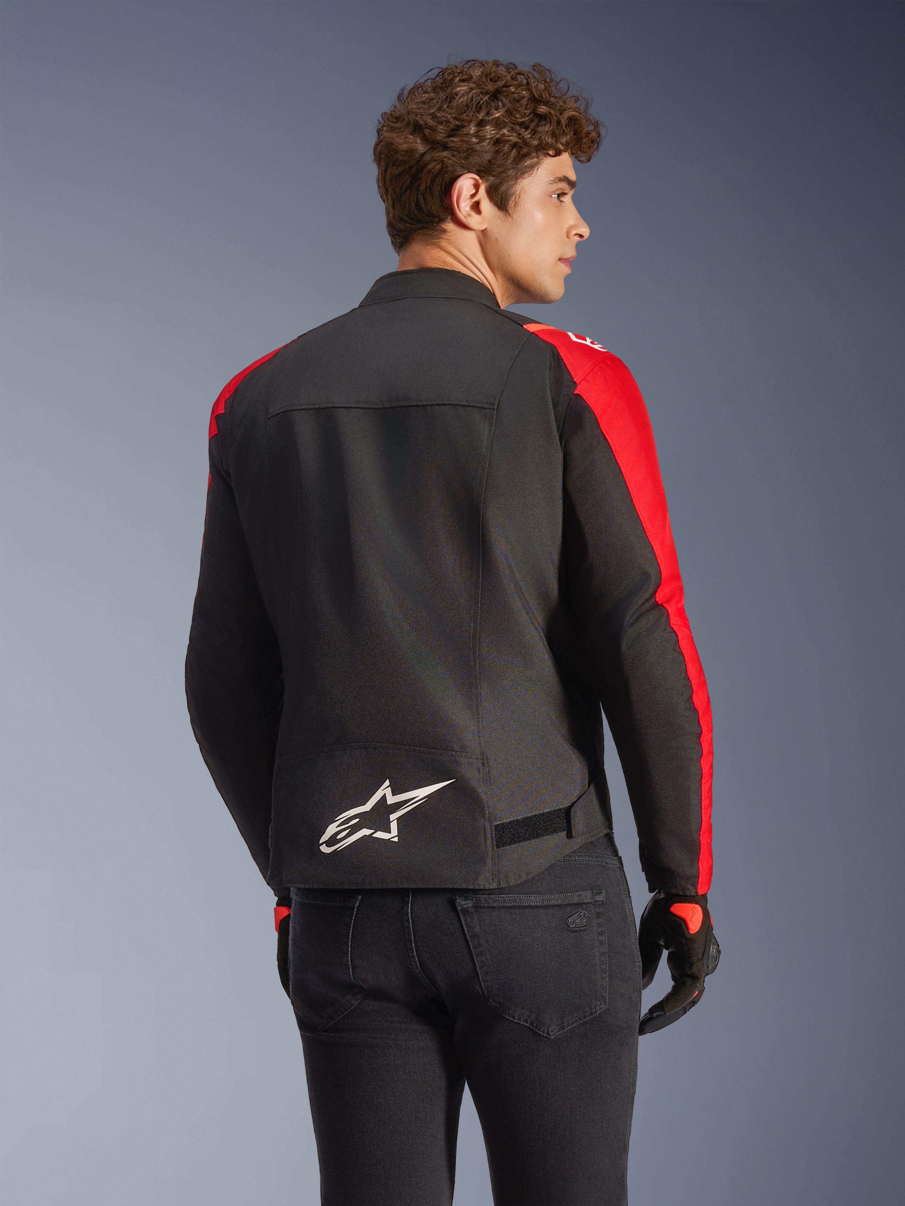 Alpinestars T-SPS V2 Waterproof Jacket in Black and Red, back view on a model, featuring a white Alpinestars logo on the lower back, vibrant red sleeve panels, and a sport-cut silhouette, worn with black gloves and jeans.