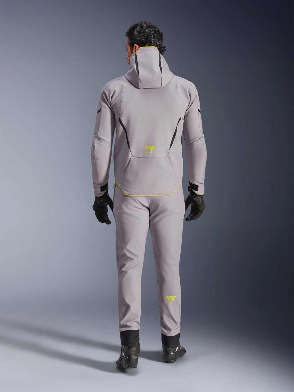 Alpinestars Aeroshell Waterproof Jacket i Mid Gray Yellow Fluo, set bagfra, båret med matchende grå bukser, sorte handsker og sorte køresko. Softshell-design med hætte, gult fluo 'ASTARS' branding og detaljer, udviklet til moderne motorcykelkørsel.