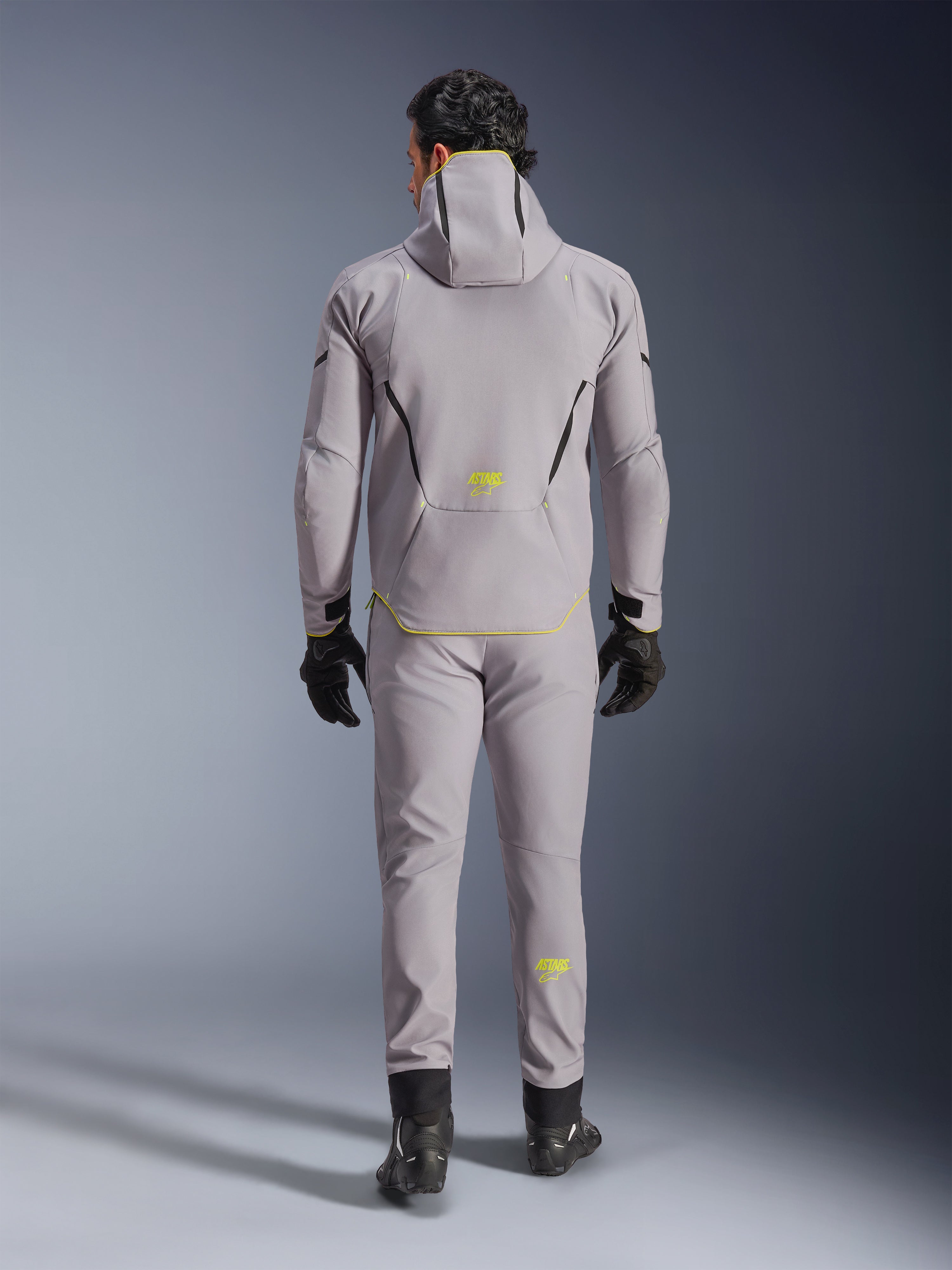 Alpinestars Aeroshell Waterproof Jacket i Mid Gray Yellow Fluo, set bagfra, båret med matchende grå bukser, sorte handsker og sorte køresko. Softshell-design med hætte, gult fluo 'ASTARS' branding og detaljer, udviklet til moderne motorcykelkørsel.