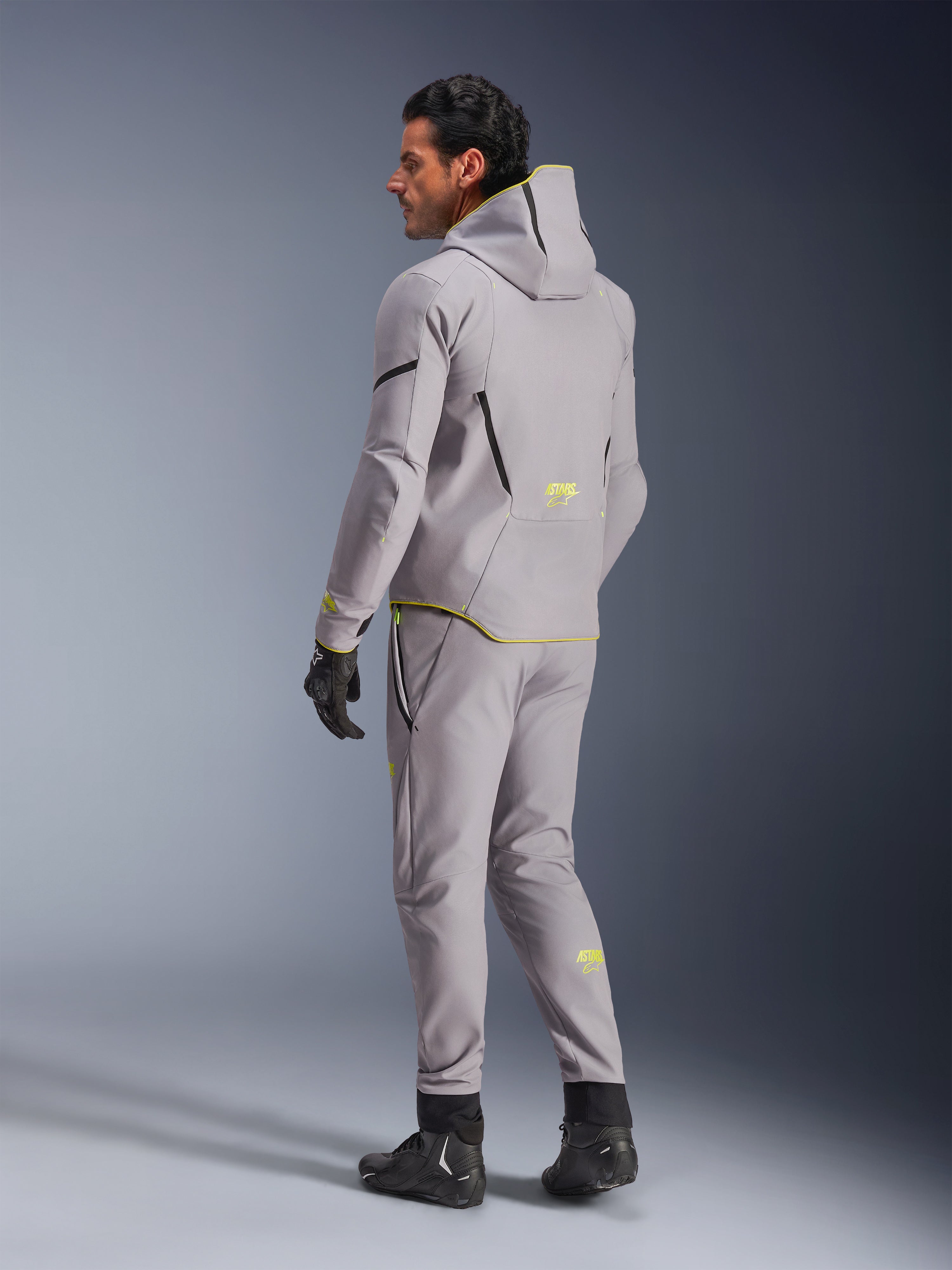 Alpinestars Aeroshell vandtæt jakke, Mid Gray Yellow Fluo, båret med matchende grå bukser, hættetrøje-design med gule detaljer og logoer, set bagfra, kombineret med sorte kørehandsker og støvler, moderne motorcykelbeklædning