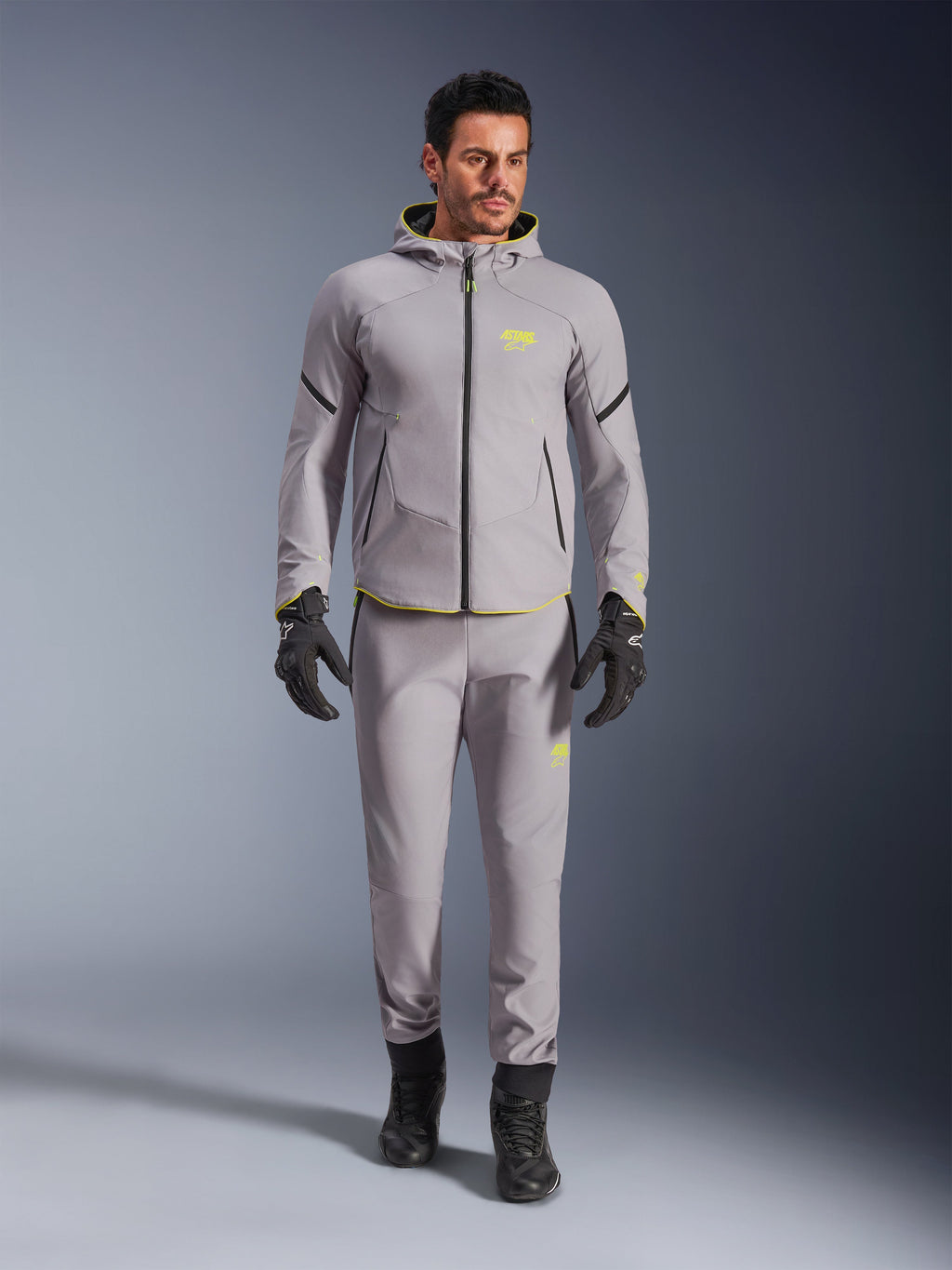 Alpinestars Aeroshell vandtæt jakke, Mid Gray Yellow Fluo, båret med matchende grå bukser, sorte handsker og støvler, med et moderne sportsinspireret hættetrøjedesign, livlige gule detaljer og atletisk pasform.