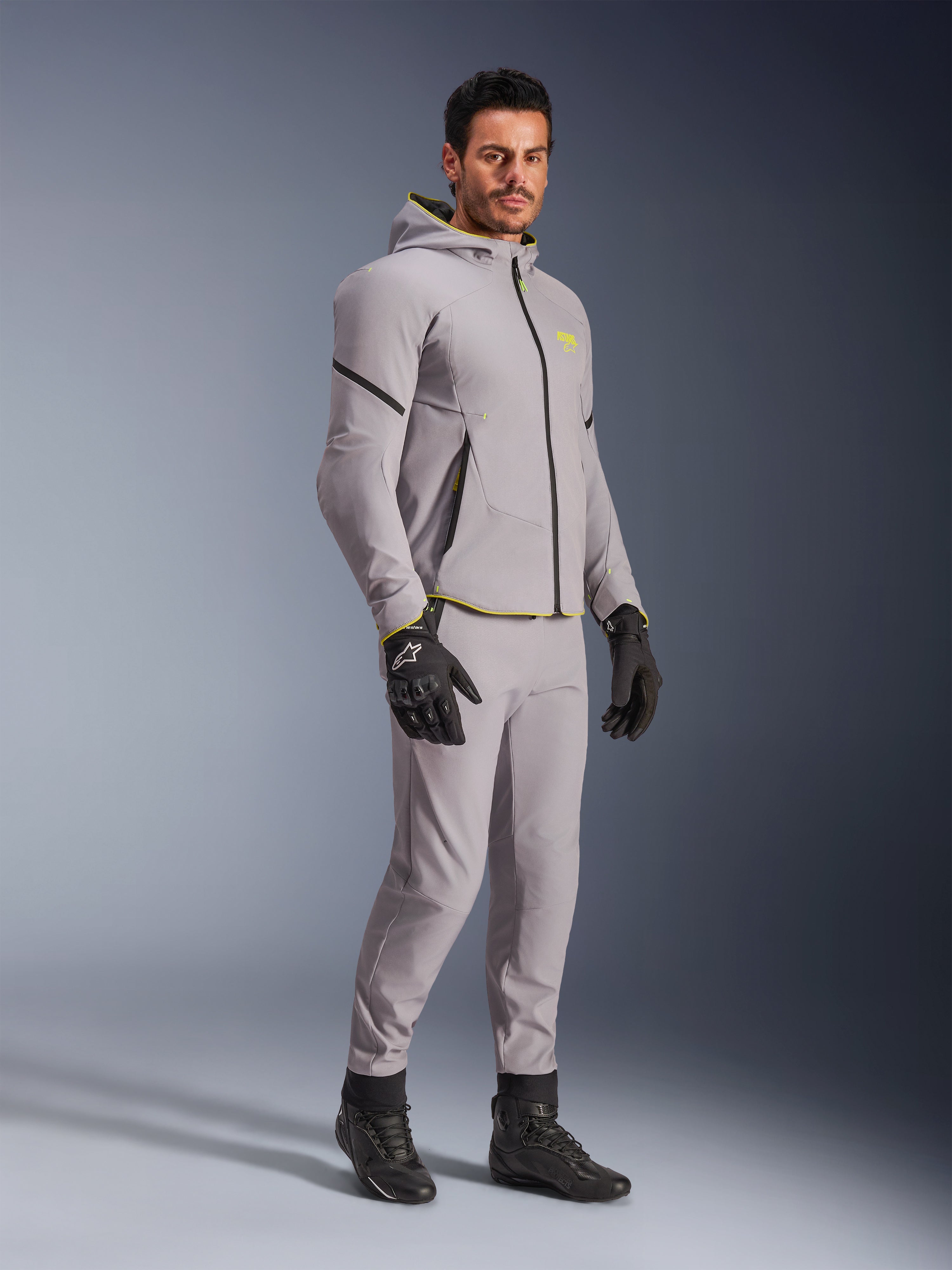Alpinestars Aeroshell vandtæt jakke, mellemgrå og neon-gul, båret af model med matchende grå bukser, sorte kørehandsker og sorte støvler, moderne motorcykeludstyr i hættetrøje-stil til casual kørsel og sportskørsel.