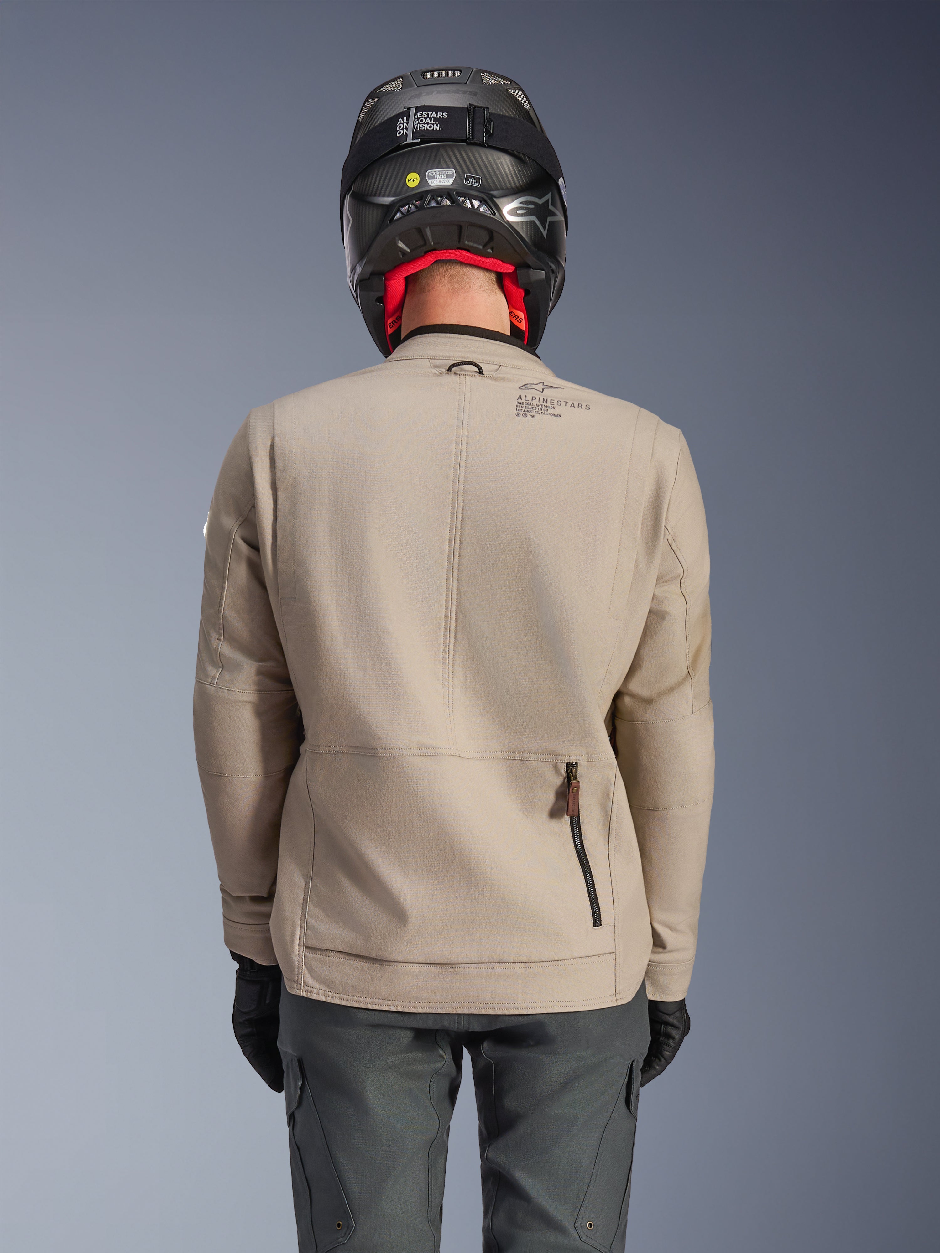 Alpinestars Borrego Drystar® jakke, adventure-motorcykeljakke, Warm Gray, set bagfra med visning af lynlås til bagrummet og Alpinestars-branding, båret med en sort hjelm og koksgrå kørebukser