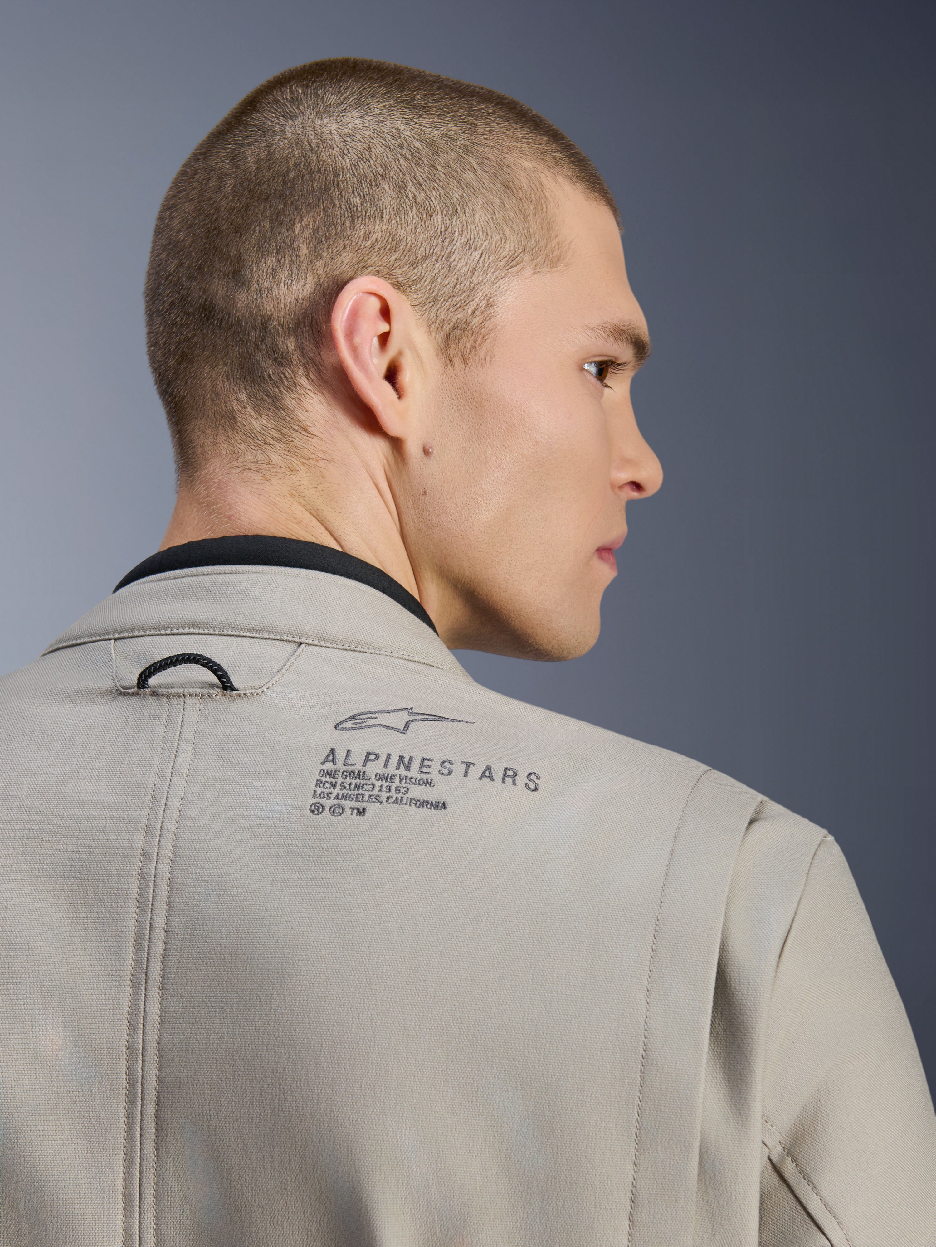 Alpinestars Borrego Drystar®-jakke i Warm Gray, nærbillede af bagskulderen, tekstureret voksbehandlet lærredsmateriale, med sort Alpinestars-logo og heritage-tekst, båret af model, adventure-motorcykeljakke