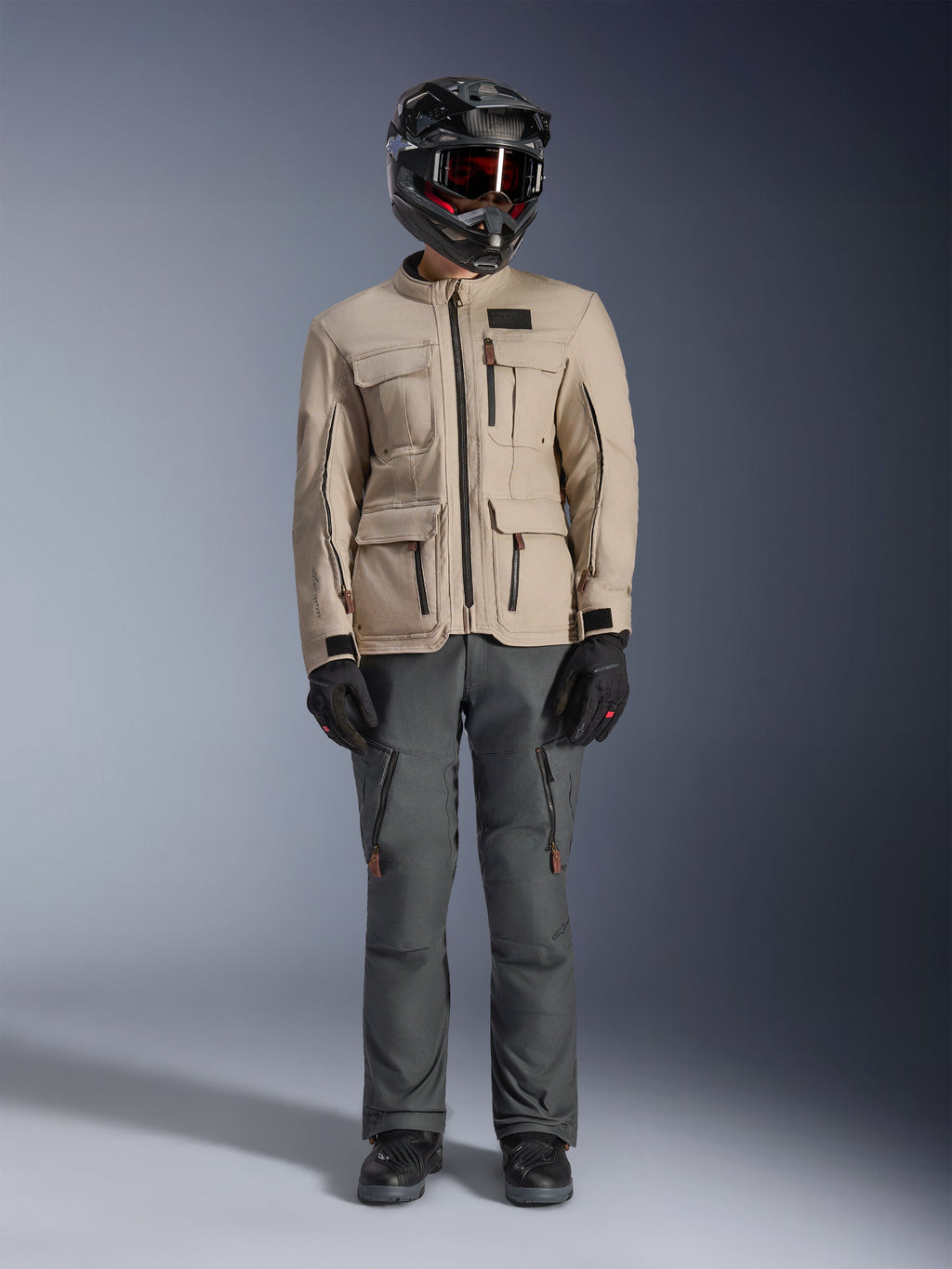 Alpinestars Borrego Drystar® jakke i Warm Gray, adventure motorcykeljakke med fire cargo-lommer foran, kombineret med mørkegrå kørebukser, sort off-road hjelm og sorte handsker, med et robust voksbehandlet lærredslook