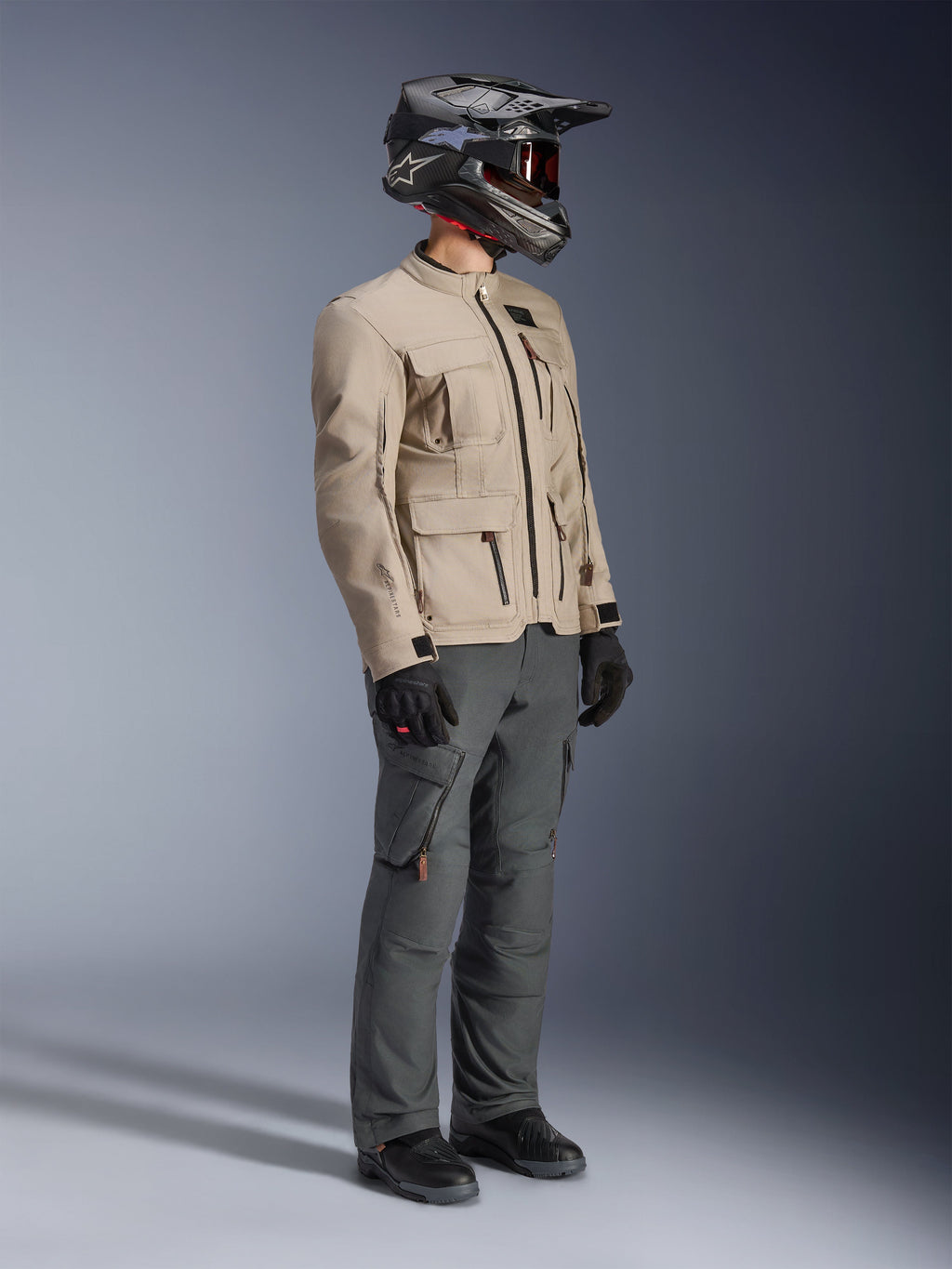 Motorcyklist iført Alpinestars Borrego Drystar® Jakke i Warm Gray, adventure motorcykeljakke med voksbehandlet lærredstekstur og praktiske lommer, stylet med mørkegrå kørebukser, kulfiberhjelm, sorte handsker og sorte støvler, fuldkropsvisning i studie af et komplet adventure touring-sæt.