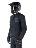 Alpinestars AMT-8 Stretch Drystar® XF jakke, sort, båret af en kører med matchende adventure-bukser og -handsker, sort motocross-hjelm og goggles, ventilationsåbninger med lynlås på bryst og arme, Alpinestars Motion Technology-branding med orange logo, førsteklasses adventure-touring-udstyr