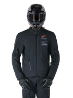 Alpinestars AMT-8 Stretch Drystar® XF-jakke, sort motorcykeljakke, med orange Alpinestars-logo og grå AMT-branding, høj krave med lynlås foran og ventilationsåbninger, bæres med sort hjelm og handsker, beskyttende adventure touring-udstyr