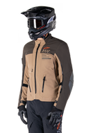 Alpinestars AMT-8 Stretch Drystar® XF Jakke, adventure motorcykeljakke, mørkebrun og mørk khaki, båret med en sort off-road hjelm og goggles, med Alpinestars Motion Technology-branding og lynlåslukkede ventilationsåbninger.