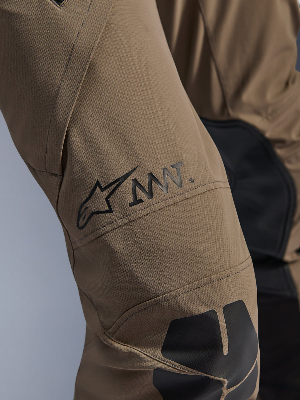 Alpinestars AMT-8 Stretch Drystar® XF bukser i Walnut Black, tætbillede af benet med det sorte AMT-logo og Alpinestars-stjerne, højtydende stræktekstil med sorte paneler, teknisk adventure-motorcykeludstyr