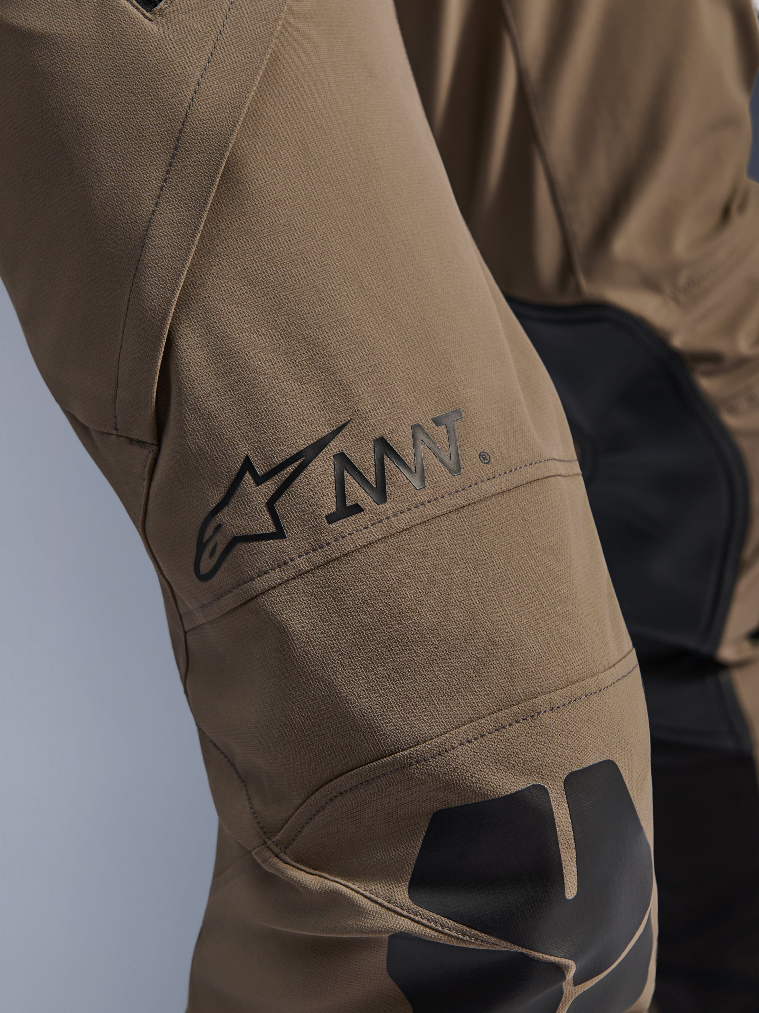 Alpinestars AMT-8 Stretch Drystar® XF bukser i Walnut Black, tætbillede af benet med det sorte AMT-logo og Alpinestars-stjerne, højtydende stræktekstil med sorte paneler, teknisk adventure-motorcykeludstyr