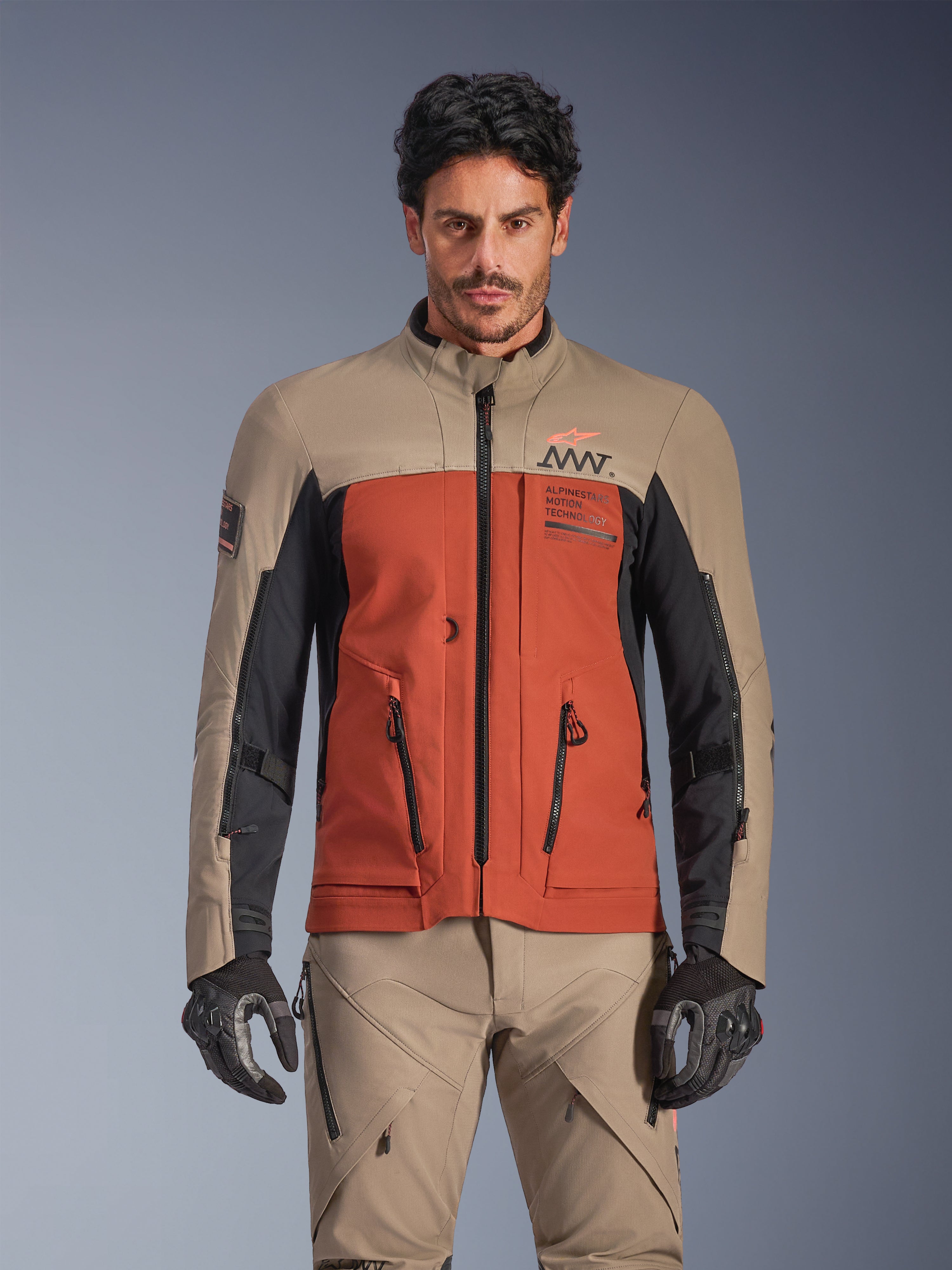 Alpinestars AMT-8 Stretch Drystar® XF Jakke, Walnut Rust Black adventure touring-jakke, båret af mandlig model med matchende bukser og handsker, med et trefarvet design i tan, rust og sort med AMT-branding på brystet, aktiv touring-pasform til motorcykelkørsel
