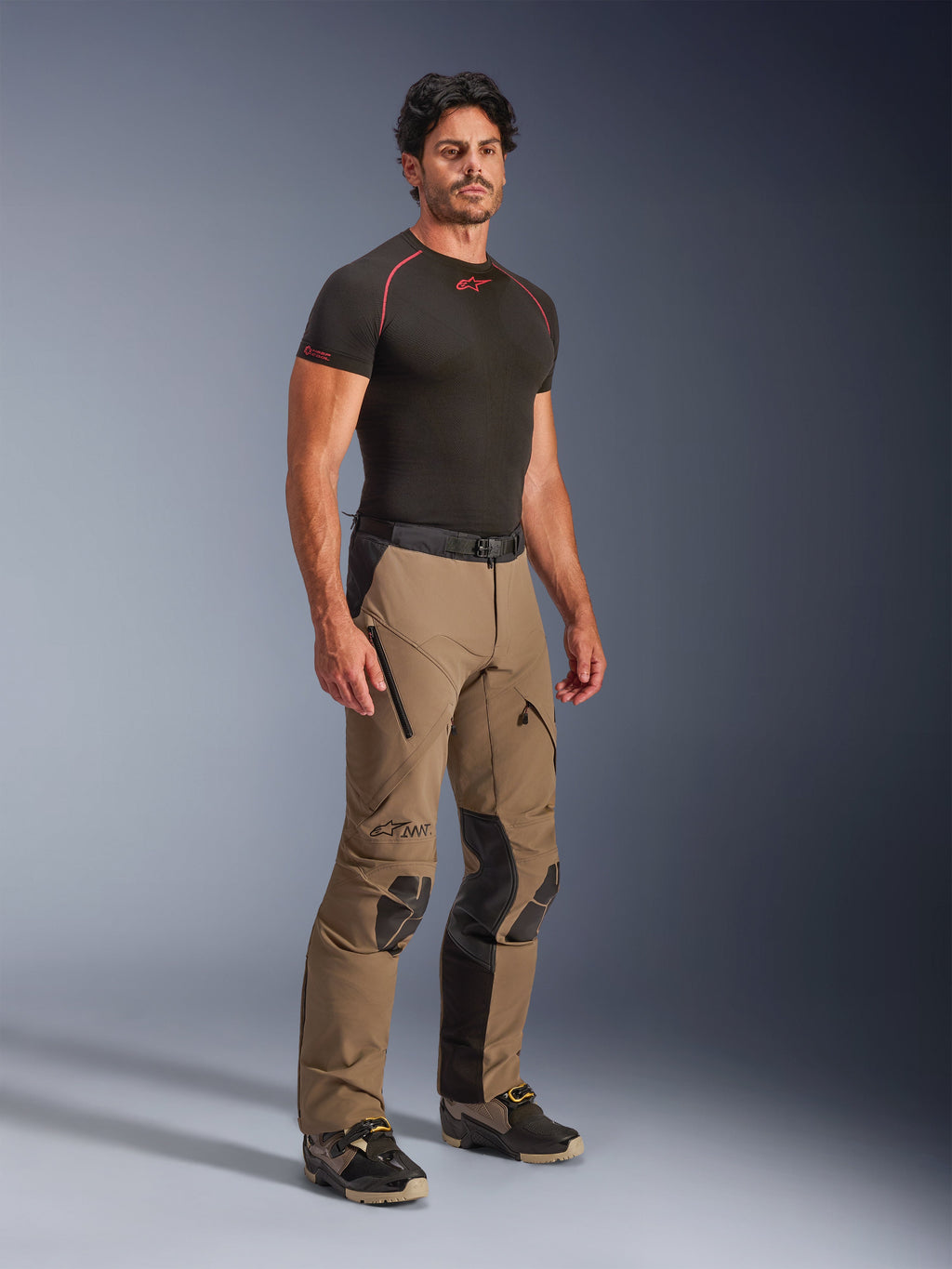 Alpinestars AMT-8 Stretch Drystar® XF bukser i Walnut Black, adventure touring motorcykelbukser båret af en model med en sort base-layer t-shirt, med strækmateriale, lårventilationsåbninger med lynlås og beskyttende knæpaneler.