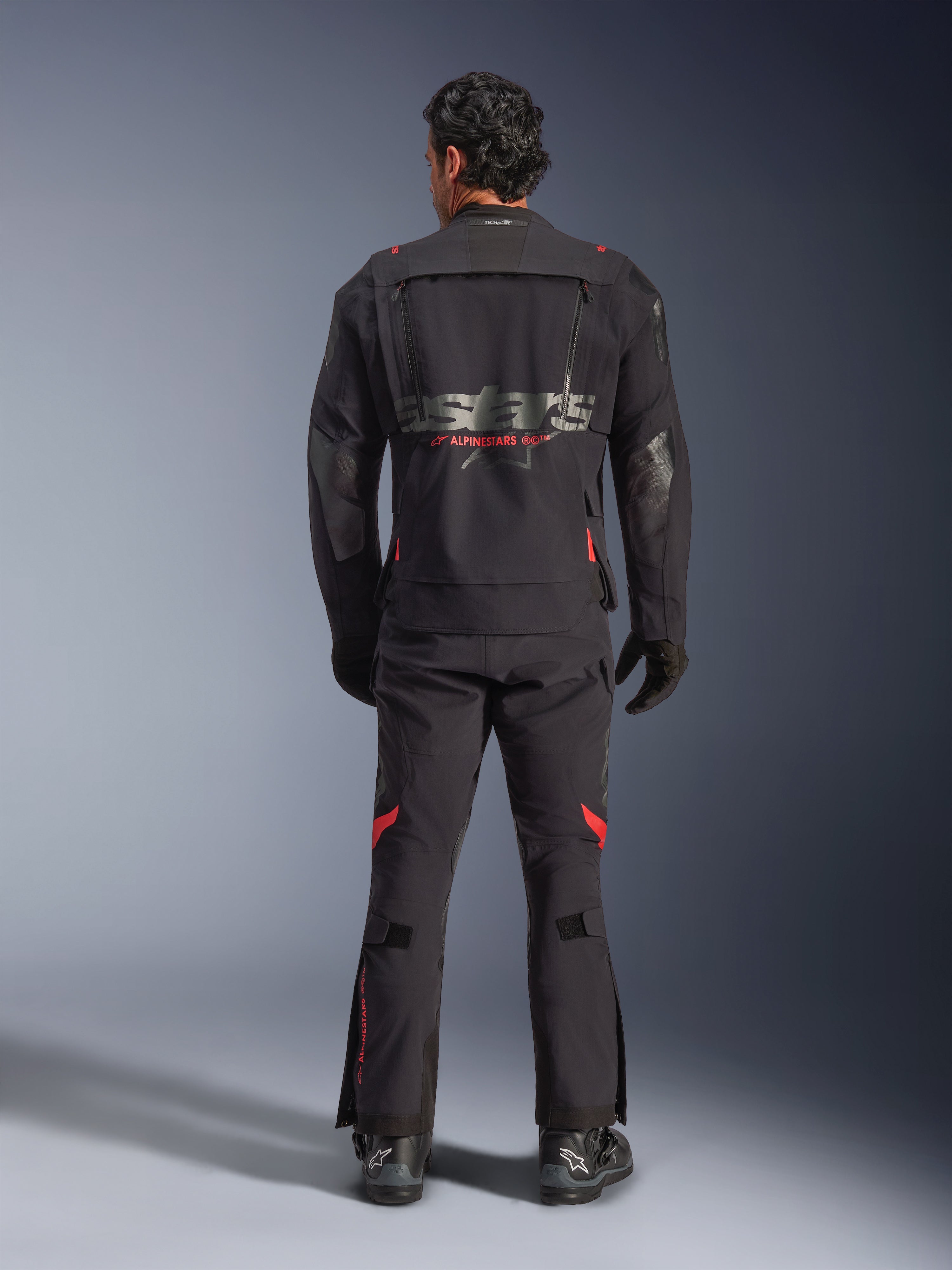 En rytter set bagfra iført Alpinestars Halo Pro Drystar® XF lamineret jakke i Black Dark Shadow, med et stort gråt astars-logo og rød Alpinestars-branding, modulære ventilationspaneler på ryggen, kombineret med matchende adventure-bukser og sorte støvler.