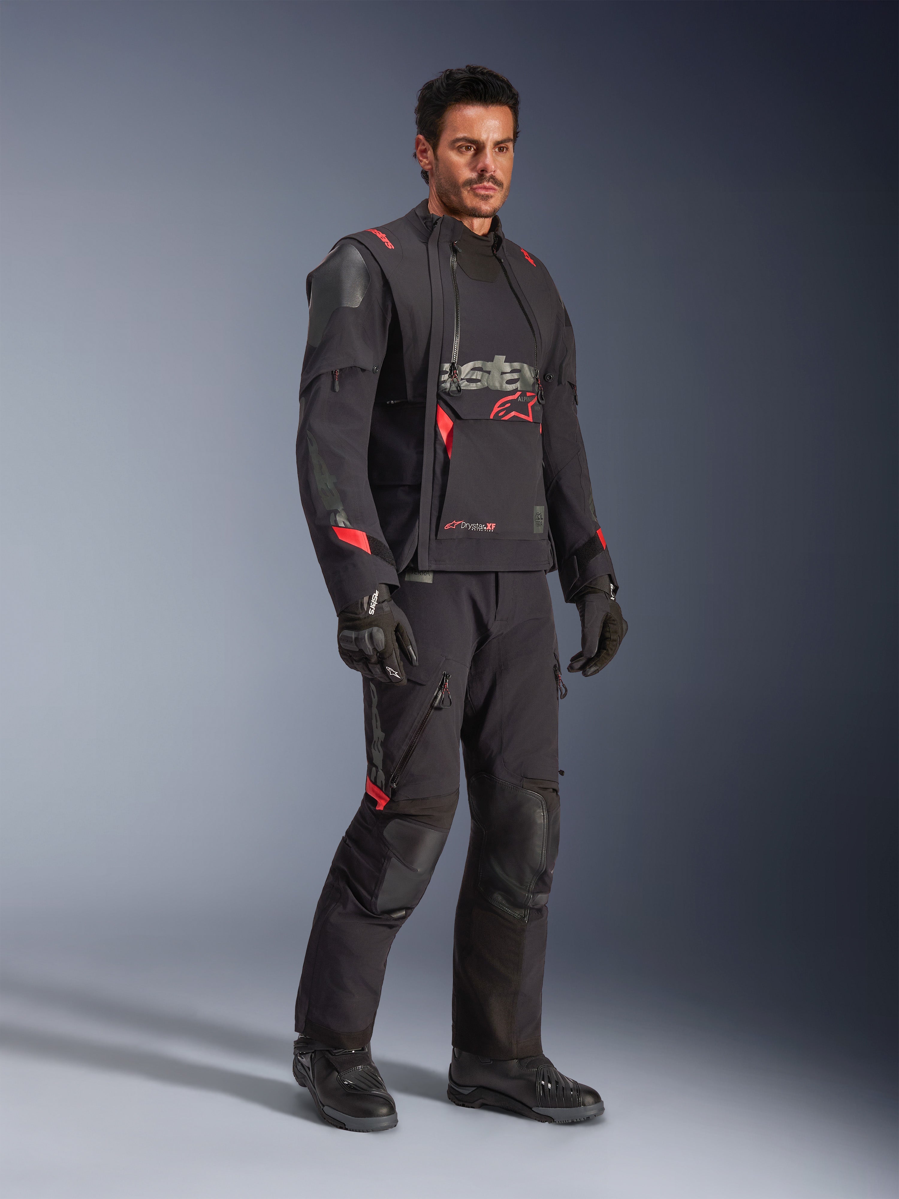Mandlig kører iført Alpinestars Halo Pro Drystar® XF lamineret jakke i Black Dark Shadow, komplet adventure-køreudstyr med matchende bukser, handsker og støvler, med modulære asymmetriske frontpaneler og røde branding-detaljer.
