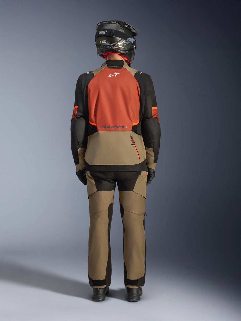 Bagfra af en kører iført Alpinestars Andes V4 Drystar®-jakke i Walnut Rust Black, kombineret med matchende valnøddefarvede adventure-bukser og en sort hjelm, der viser jakkens rustfarvede rygpanel, store baglomme og reflekterende logoer.