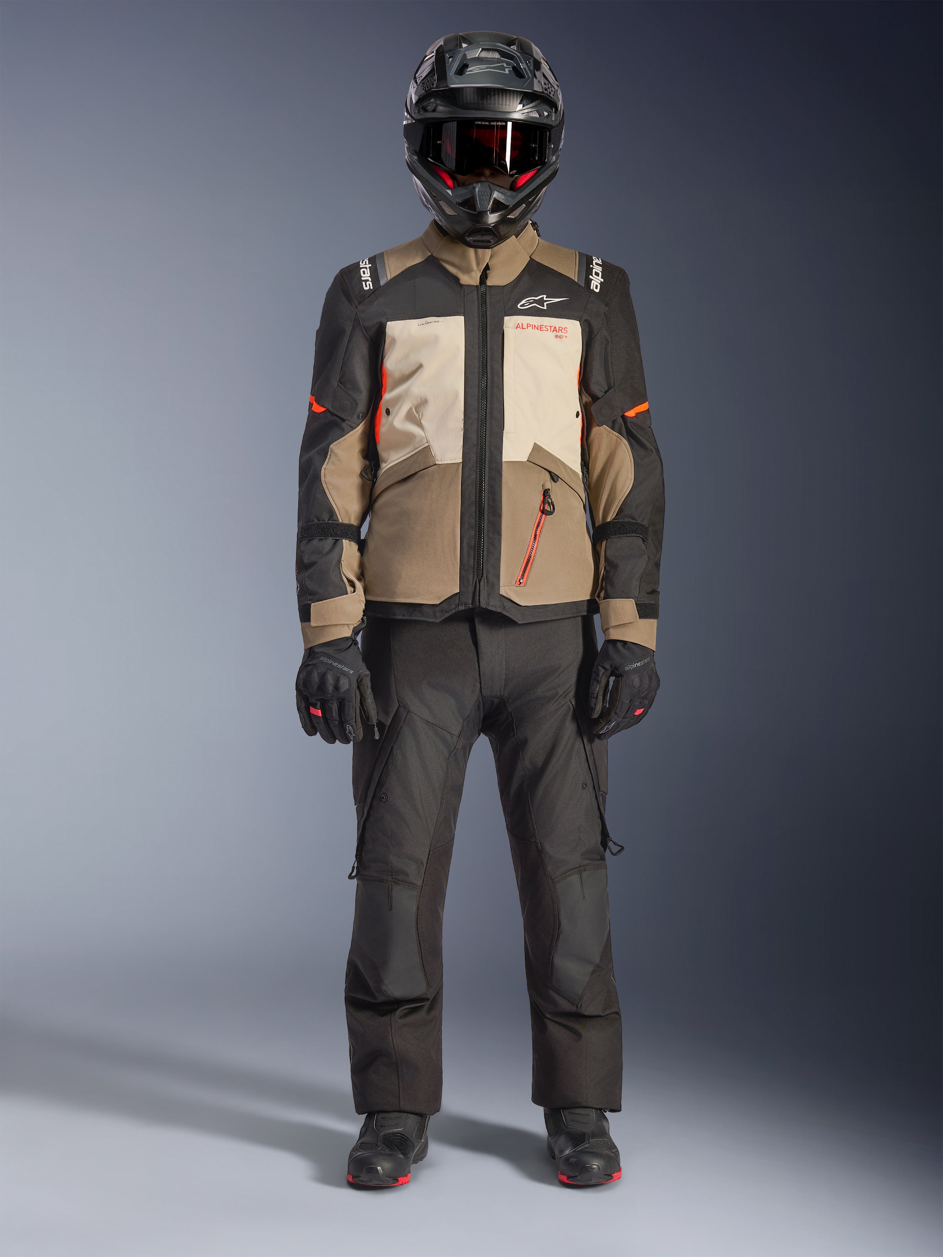 Alpinestars Andes V4 Drystar® Jakke, Walnut Mastic Black, adventure touring motorcykeljakke med tan- og cremefarvede paneler og orange detaljer, båret af en rytter med sort adventure-hjelm, sorte motorcykelbukser og sorte støvler, fuldkropsvisning i studie.