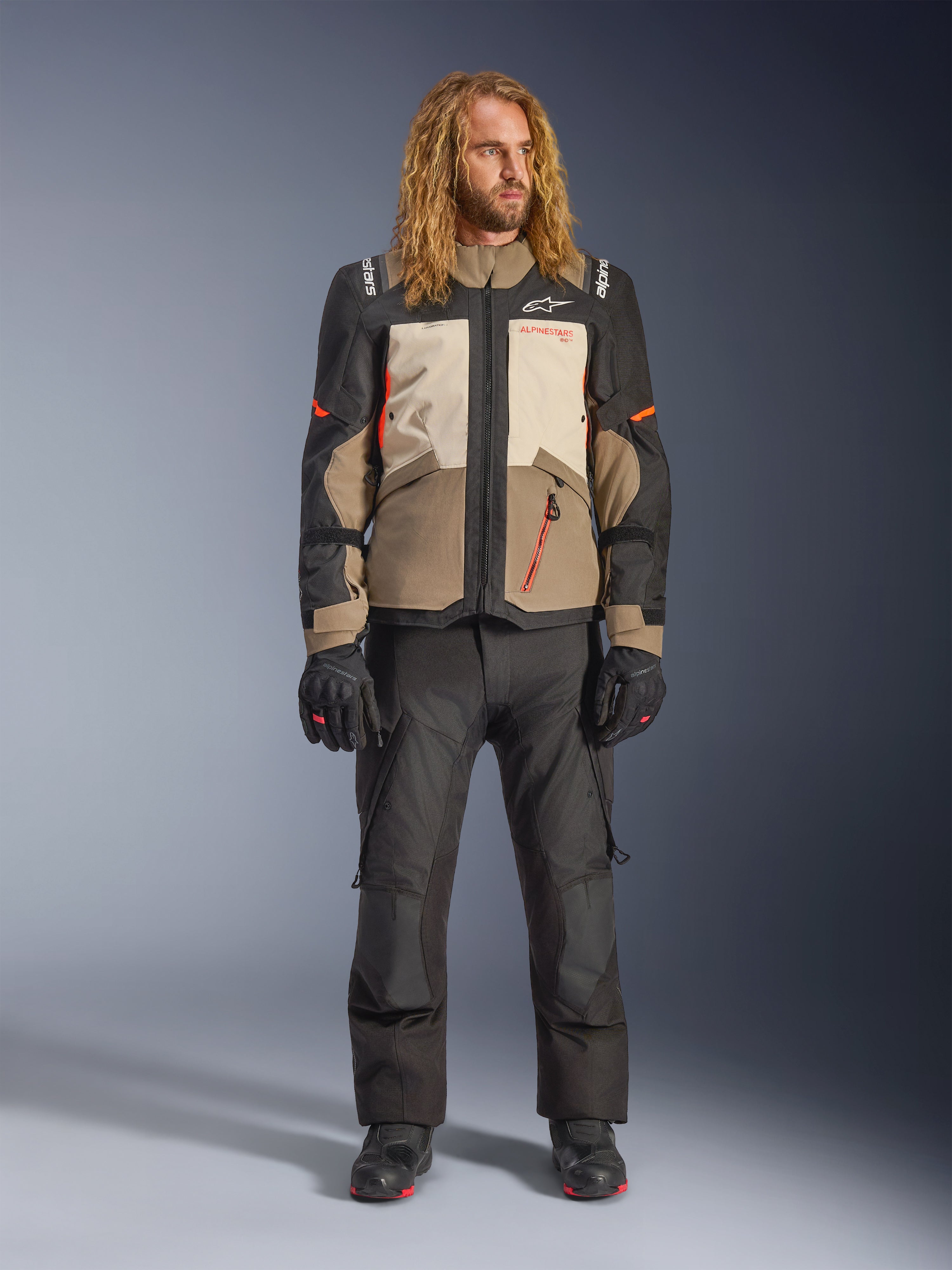 Alpinestars Andes V4 Drystar®-jakke, Walnut Mastic Black, båret med sorte motorcykelbukser og handsker, med et multi-panel adventure-design med orange detaljer og Alpinestars-branding, teknisk tekstiludstyr til kørsel i al slags vejr
