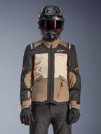 Alpinestars Andes V4 Drystar® Jakke, Walnut Mastic Black, adventure-touringjakke med valnøddebrune og mastic-cremefarvede paneler, sorte ærmer og klare orange detaljer, båret med en sort motorcykelhjelm, mørke goggles og sorte handsker