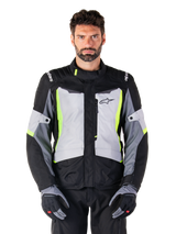 Alpinestars ST-1 Waterproof Jacket, adventure touring motorcykeljakke, Ice Gray Black og Yellow Fluo, båret af en mand med matchende handsker og sorte bukser, med neongule detaljer på brystet og kraven, Alpinestars-logo på brystet