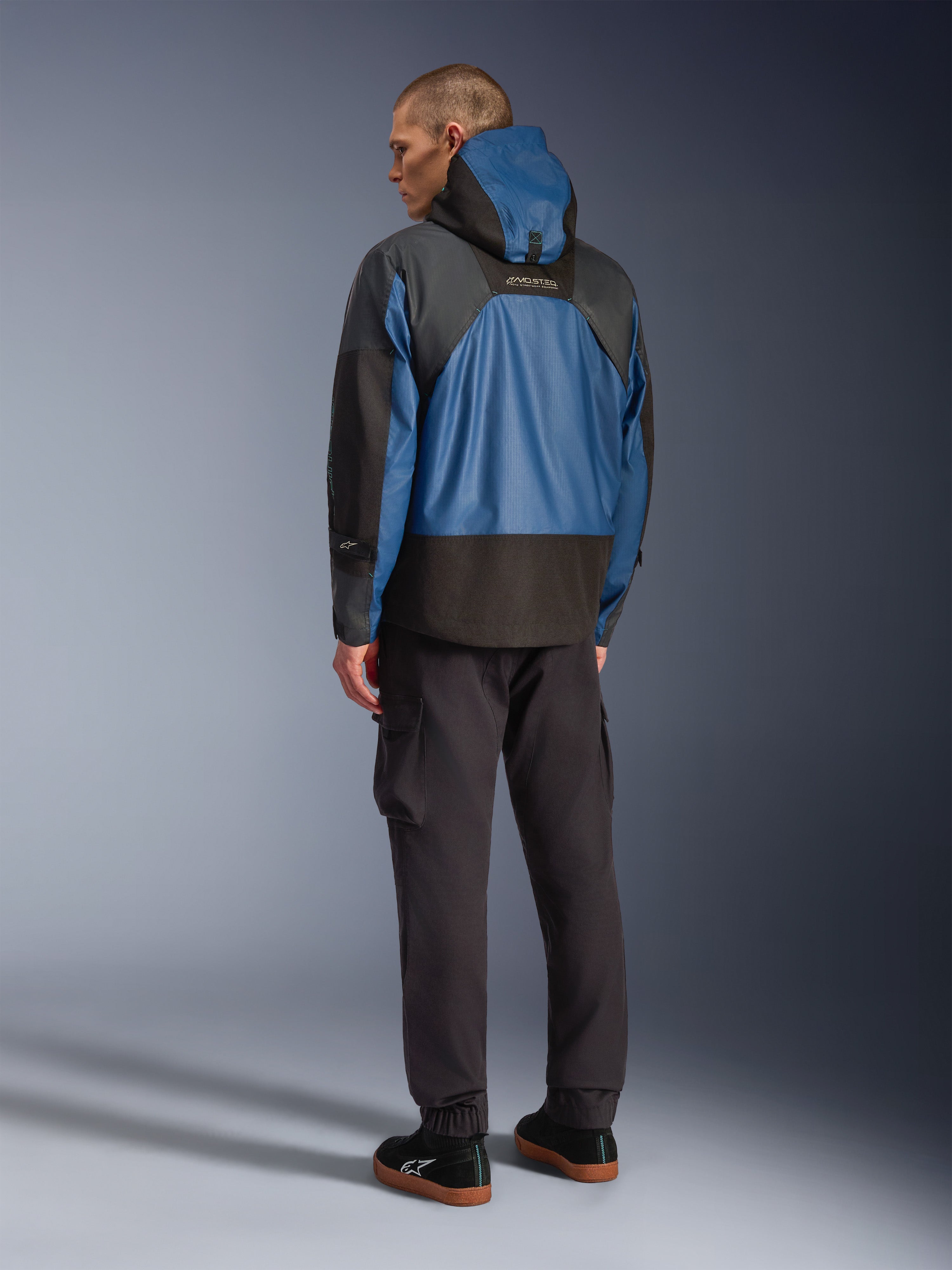 Set bagfra af en mandlig model iført Alpinestars Mospher Waterproof Jacket i Black og Dark Petrol, med et design med hætte og taktisk ripstop-konstruktion, kombineret med sorte cargobukser og sneakers.