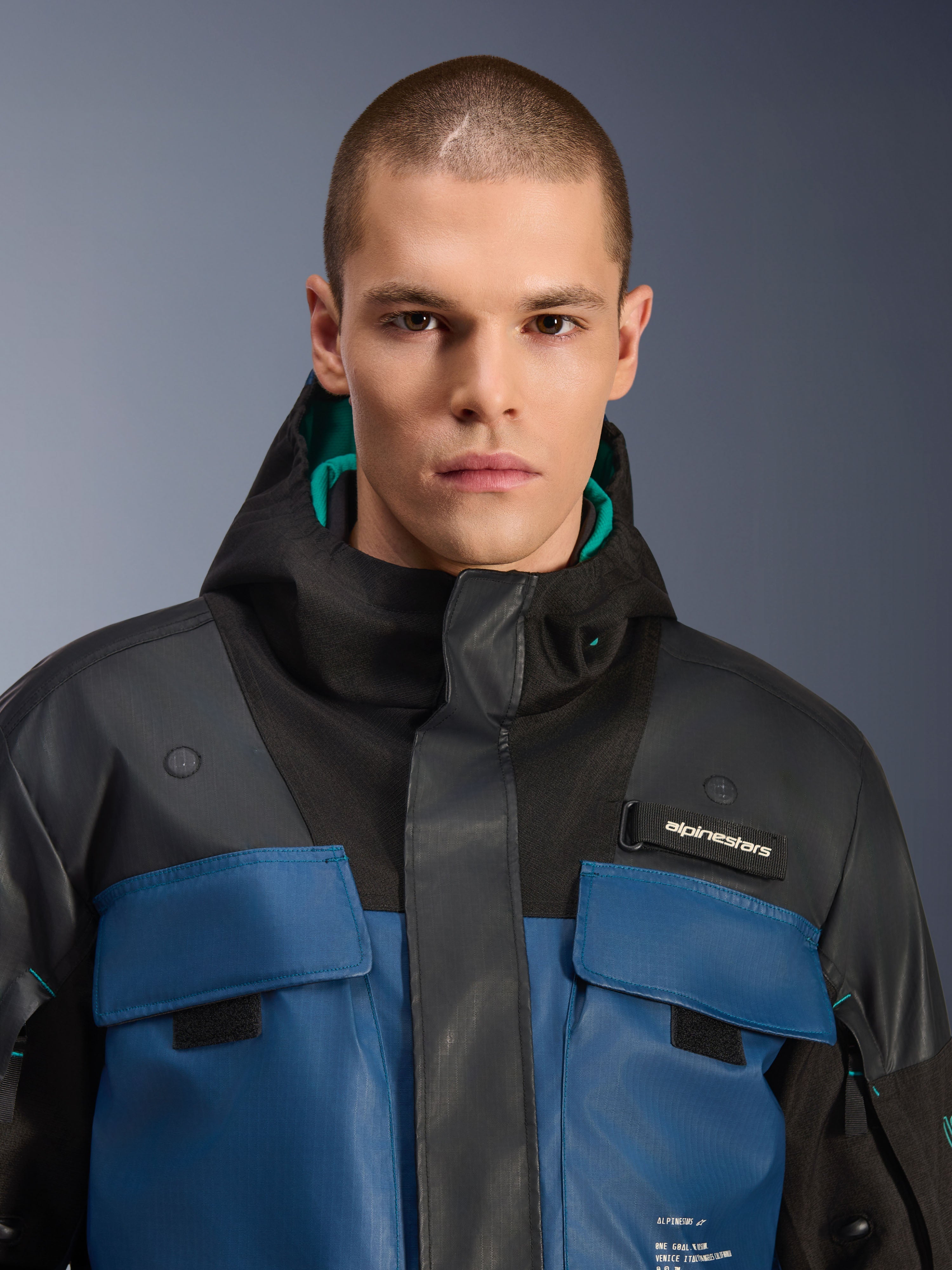 Alpinestars Mospher Waterproof Jacket, sort og mørk petrol, nærbillede af mandlig model iført den utility-inspirerede parka, med mørkeblå brystlommer, sort hætte med teal-farvet for og Alpinestars-branding på brystet.