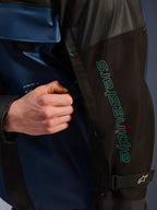 Alpinestars Mospher vandtæt jakke i Black Dark Petrol, nærbillede af ærmet med petroleumsblå Alpinestars-branding, en hånd der justerer ventilationslynlåsen, slidstærkt ripstop-stof og en justerbar armrem