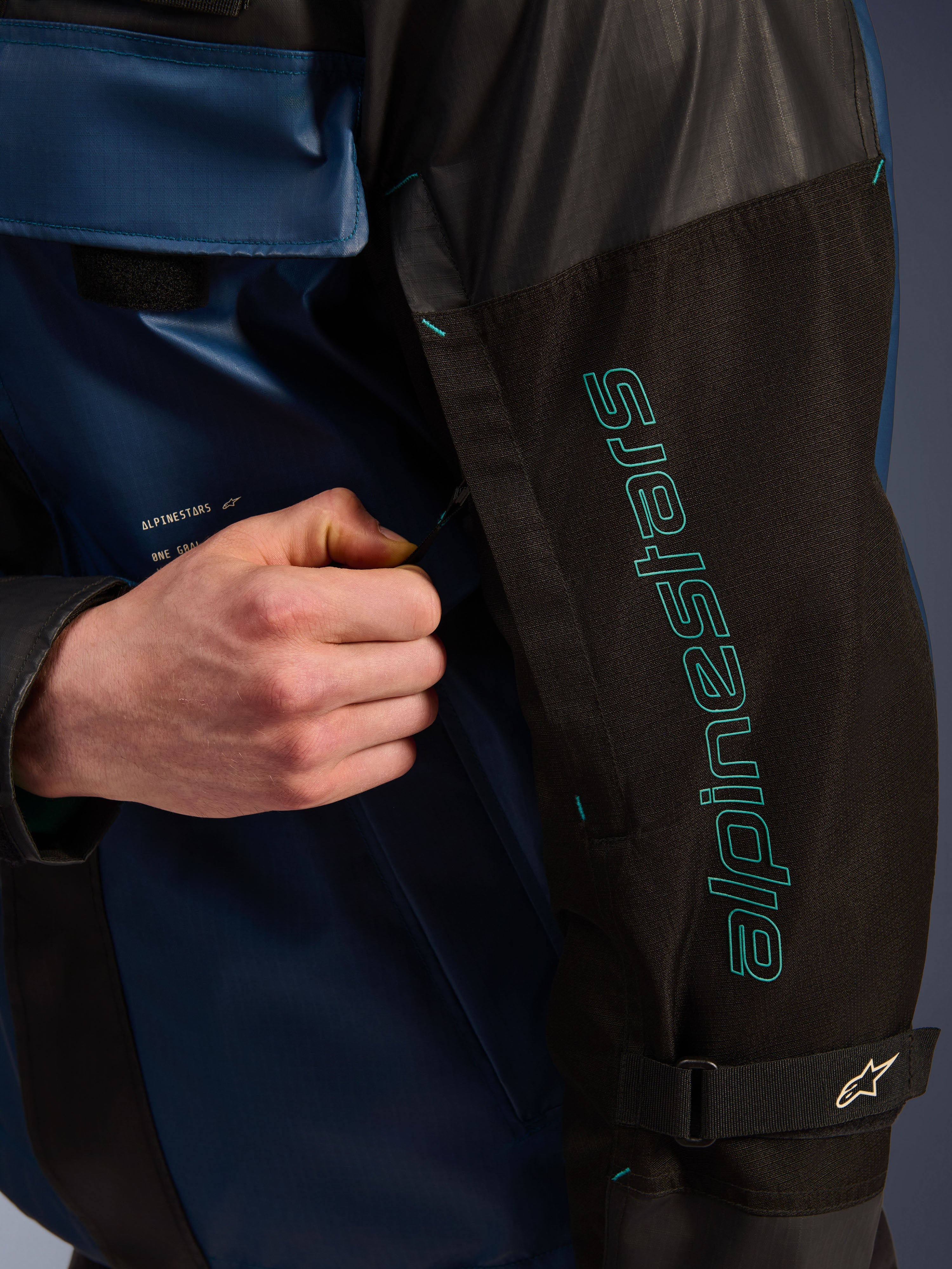 Alpinestars Mospher vandtæt jakke i Black Dark Petrol, nærbillede af ærmet med petroleumsblå Alpinestars-branding, en hånd der justerer ventilationslynlåsen, slidstærkt ripstop-stof og en justerbar armrem