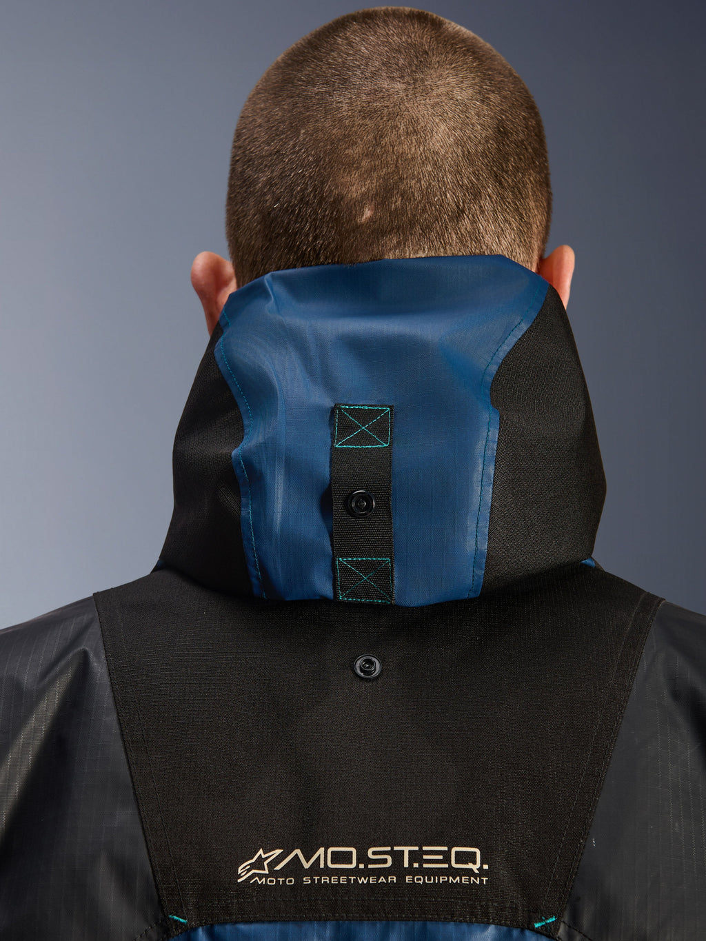 Alpinestars Mospher Waterproof-jakke, detalje set bagfra der viser hætteforankringssystemet, sort og Dark Petrol Blue, med MO.ST.EQ. Moto Streetwear Equipment-branding, slidstærkt ripstop-tekstilmateriale, teknisk pendlermotorcykeludstyr
