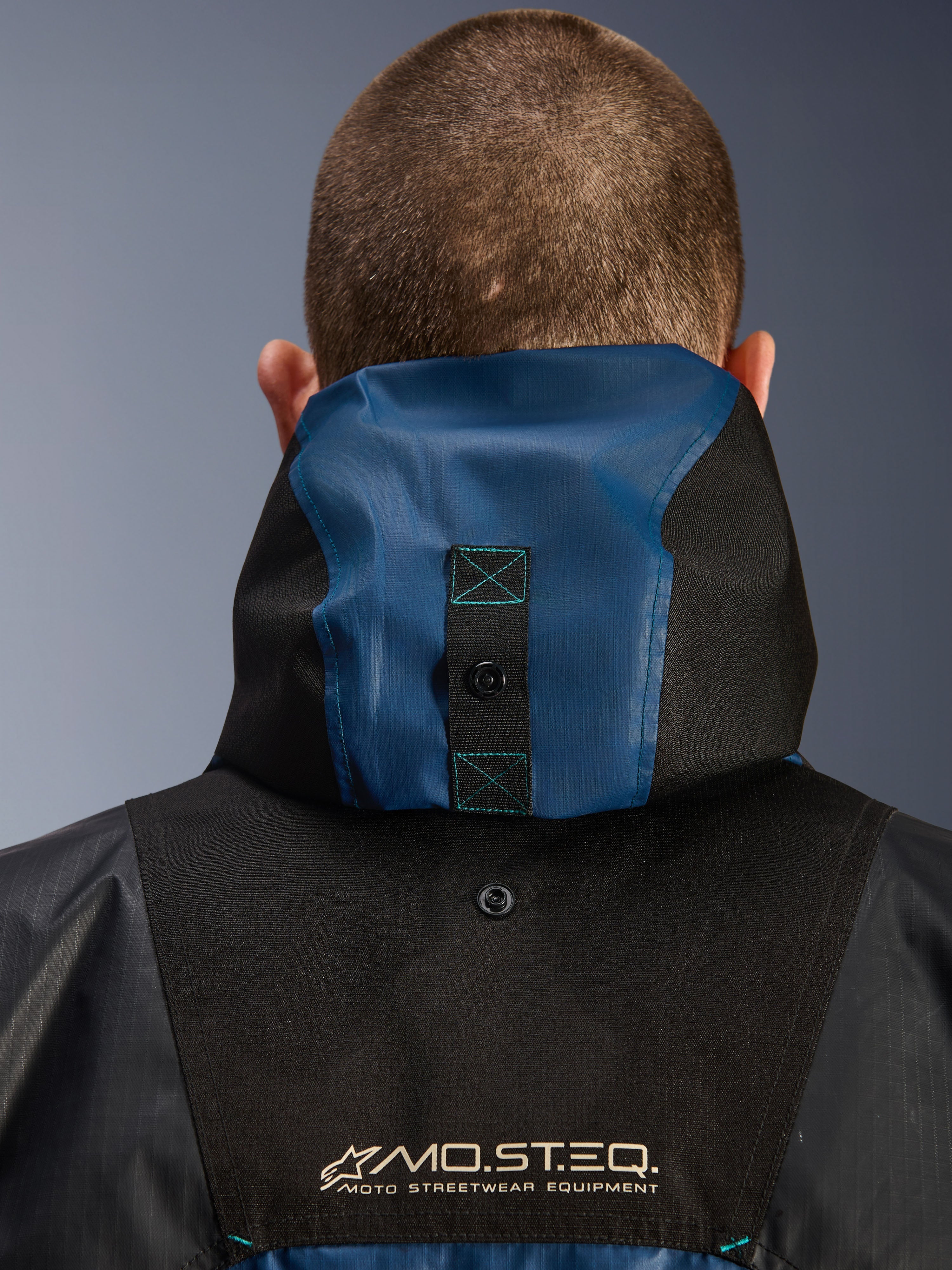 Alpinestars Mospher Waterproof-jakke, detalje set bagfra der viser hætteforankringssystemet, sort og Dark Petrol Blue, med MO.ST.EQ. Moto Streetwear Equipment-branding, slidstærkt ripstop-tekstilmateriale, teknisk pendlermotorcykeludstyr
