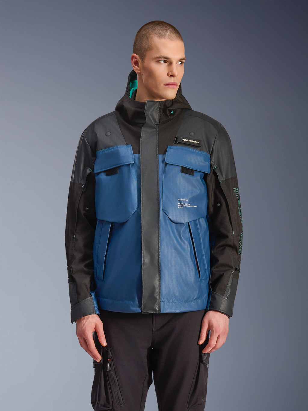 Alpinestars Mospher vandtæt jakke, urban motorcykelparka, sort og mørk petrolblå, store taktiske brystlommer, høj krave med hætte, teknisk ripstop-tekstur, set forfra på model