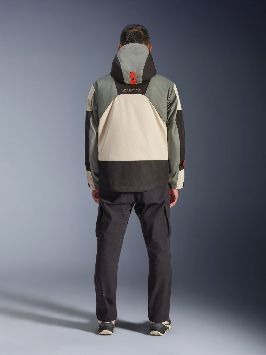 Set bagfra af en kører iført Alpinestars Mospher Waterproof Jacket i Fur Gray Black Ice, taktisk inspireret parka med grå, sorte og cremefarvede farveblokke, har en hætte med orange remdetalje, parret med sorte cargobukser.