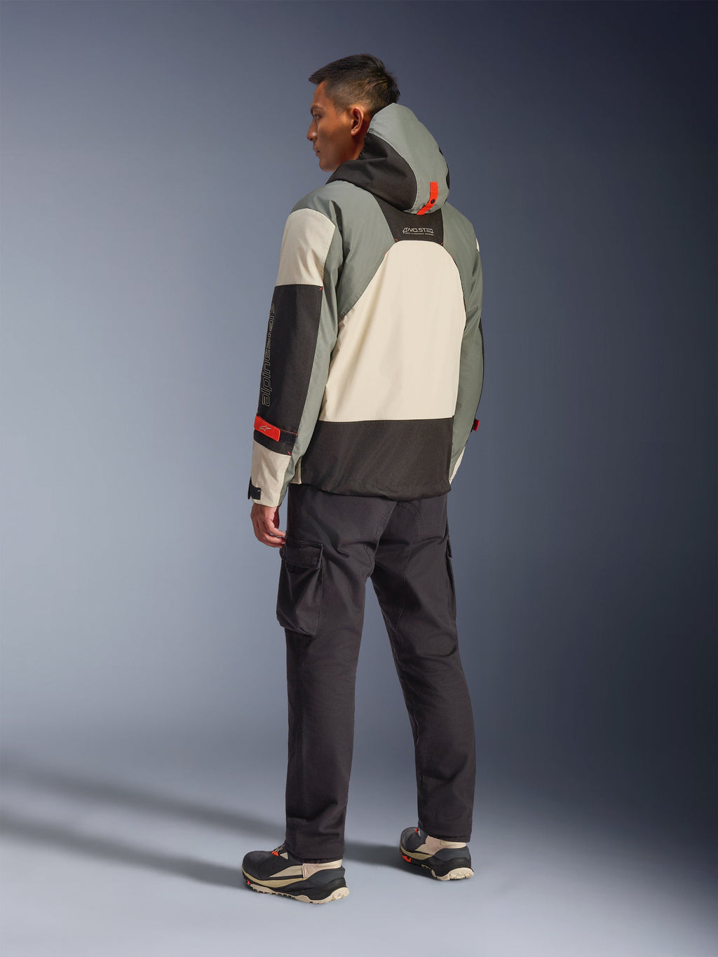 Alpinestars Mospher vandtæt jakke i Fur Gray Black Ice, set bagfra på en model iført en taktisk motorcykelparka med cremefarvede, grå og sorte paneler, med røde detaljer og en hætte, kombineret med sorte cargo-bukser og tekniske støvler