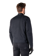Alpinestars Clayton WR Jakke i sort, trekvart visning bagfra på en mandlig model, urban motorcykeljakke med quiltet polstring på skuldre og albuer, tekniske syninger og reflekterende logo på kraven, stylet med sorte bukser og handsker.