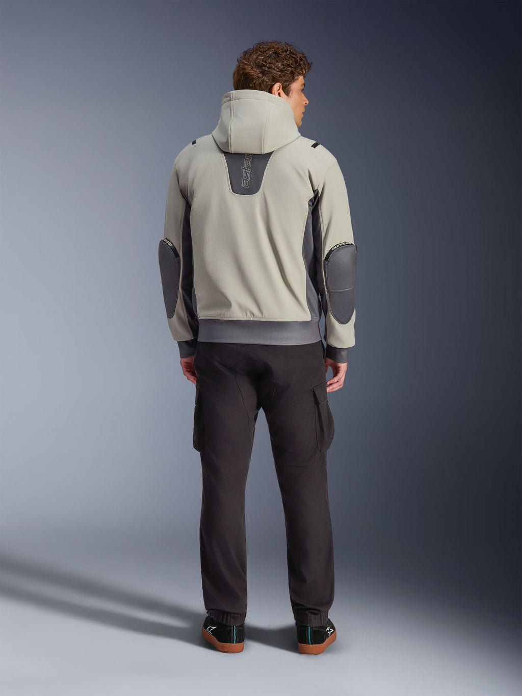 Alpinestars Moblast Waterproof Jacket, Iron Gray Dark Sage, set bagfra af en mandlig kører iført motorcykeljakke med hætte, lysegrå og mørk salviegrøn farvekombination, lodret 'astars' logo på den øverste del af ryggen, mørkegrå albuelapper, stylet med mørke cargo-bukser for et urbant, moderne look
