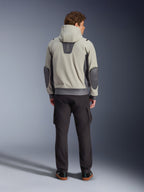 Alpinestars Moblast Waterproof Jacket, Iron Gray Dark Sage, set bagfra af en mandlig kører iført motorcykeljakke med hætte, lysegrå og mørk salviegrøn farvekombination, lodret 'astars' logo på den øverste del af ryggen, mørkegrå albuelapper, stylet med mørke cargo-bukser for et urbant, moderne look