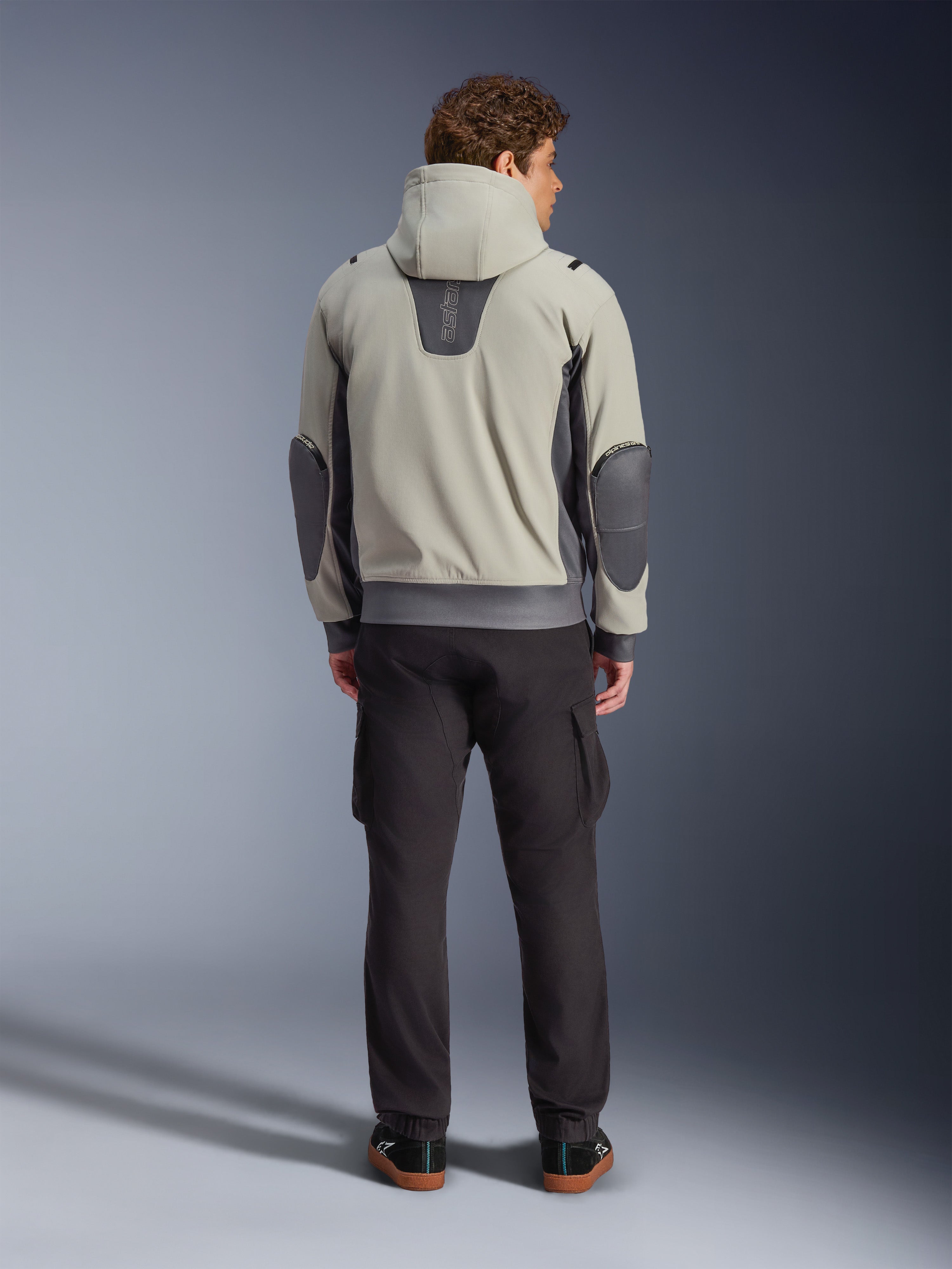 Alpinestars Moblast Waterproof Jacket, Iron Gray Dark Sage, set bagfra af en mandlig kører iført motorcykeljakke med hætte, lysegrå og mørk salviegrøn farvekombination, lodret 'astars' logo på den øverste del af ryggen, mørkegrå albuelapper, stylet med mørke cargo-bukser for et urbant, moderne look