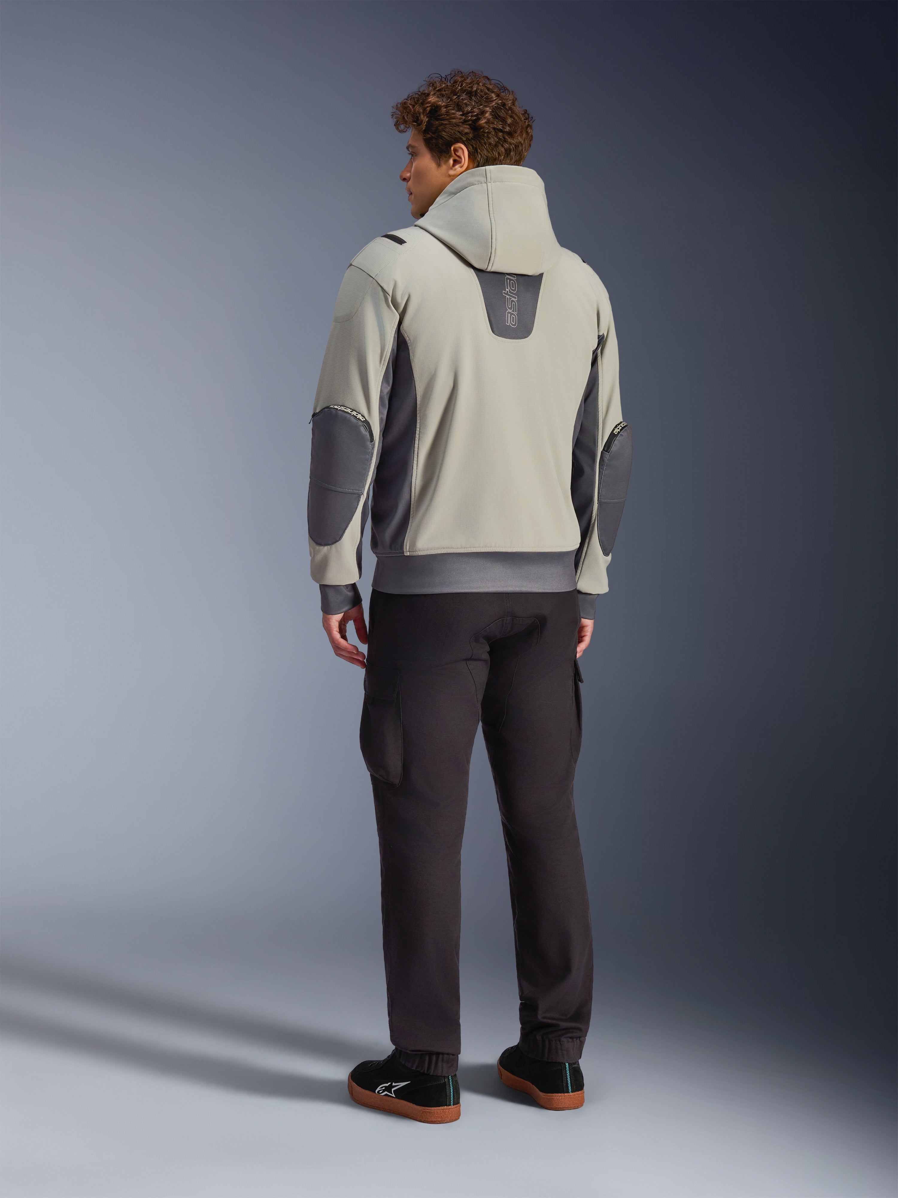 Mandlig model set bagfra iført Alpinestars Moblast Waterproof Jacket i Iron Gray Dark Sage, urban motorcykeljakke med hætte, mørkegrå sidepaneler og forstærkede albuelapper, kombineret med mørke kørebukser og sorte sneakers, moderne minimalistisk æstetik til urban pendling