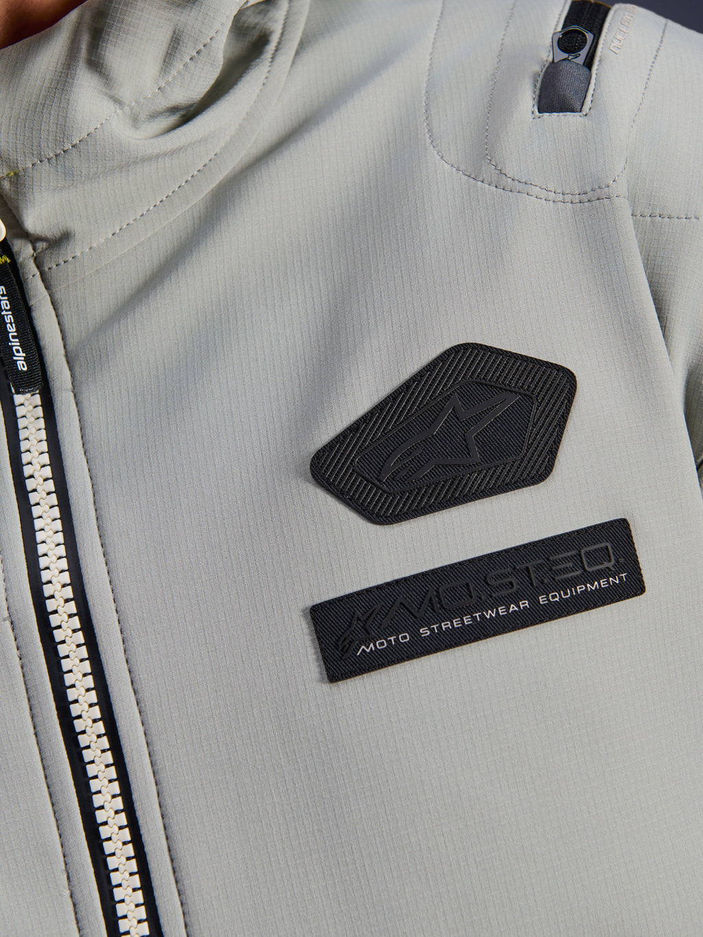 Alpinestars Moblast vandtæt jakke, Iron Gray, nærbillede af brystmærker med Alpinestars-logo og MO.ST.EQ. Moto Streetwear Equipment-branding, teknisk ripstop-materiale med kraftig lynlås foran og detalje på skulderlomme