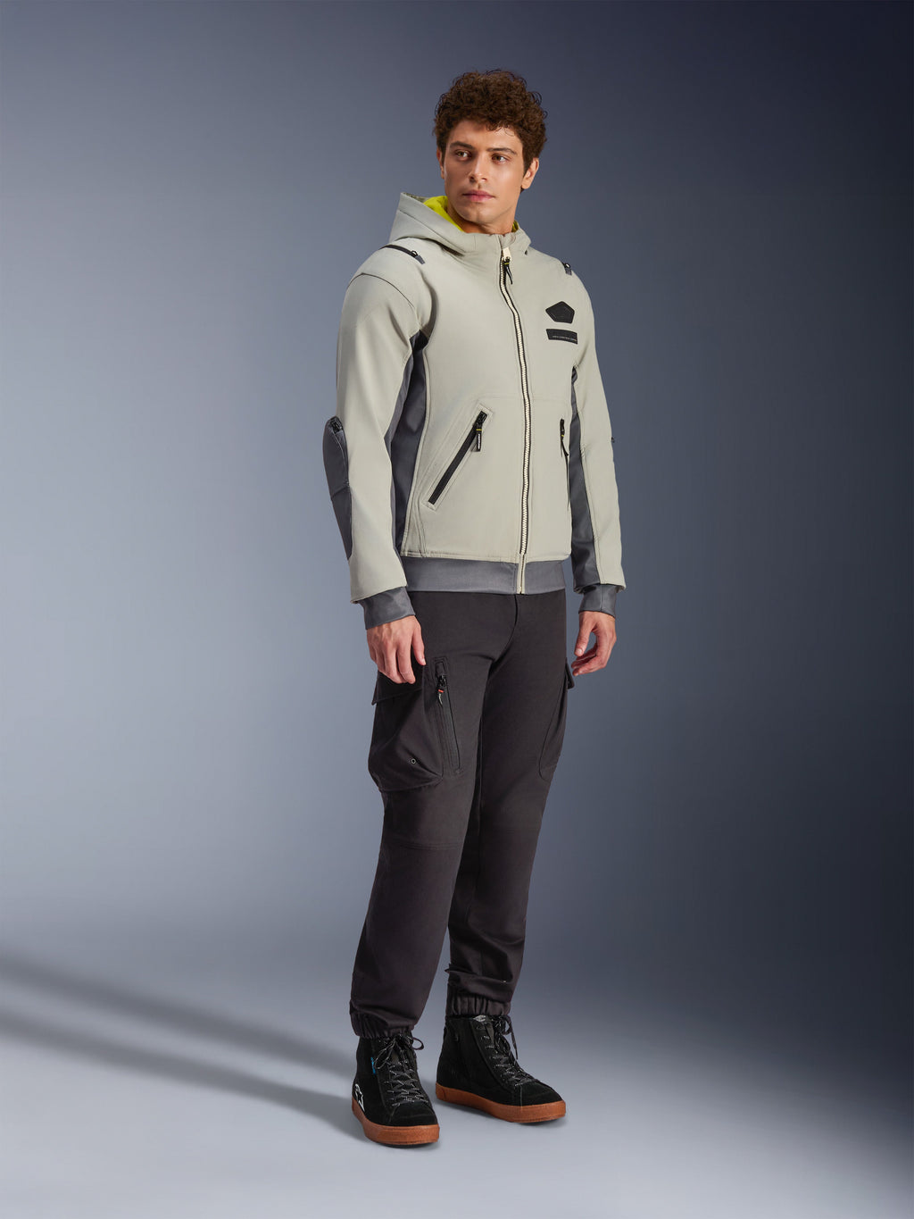 Alpinestars Moblast vandtæt jakke, Iron Gray og Dark Sage, urban motorcykeljakke med hættetrøjedesign, båret af en model med mørke kørebukser og sorte støvler, minimalistisk cyber-tech-æstetik med gult indvendigt for i hætten og vandtætte lynlåse